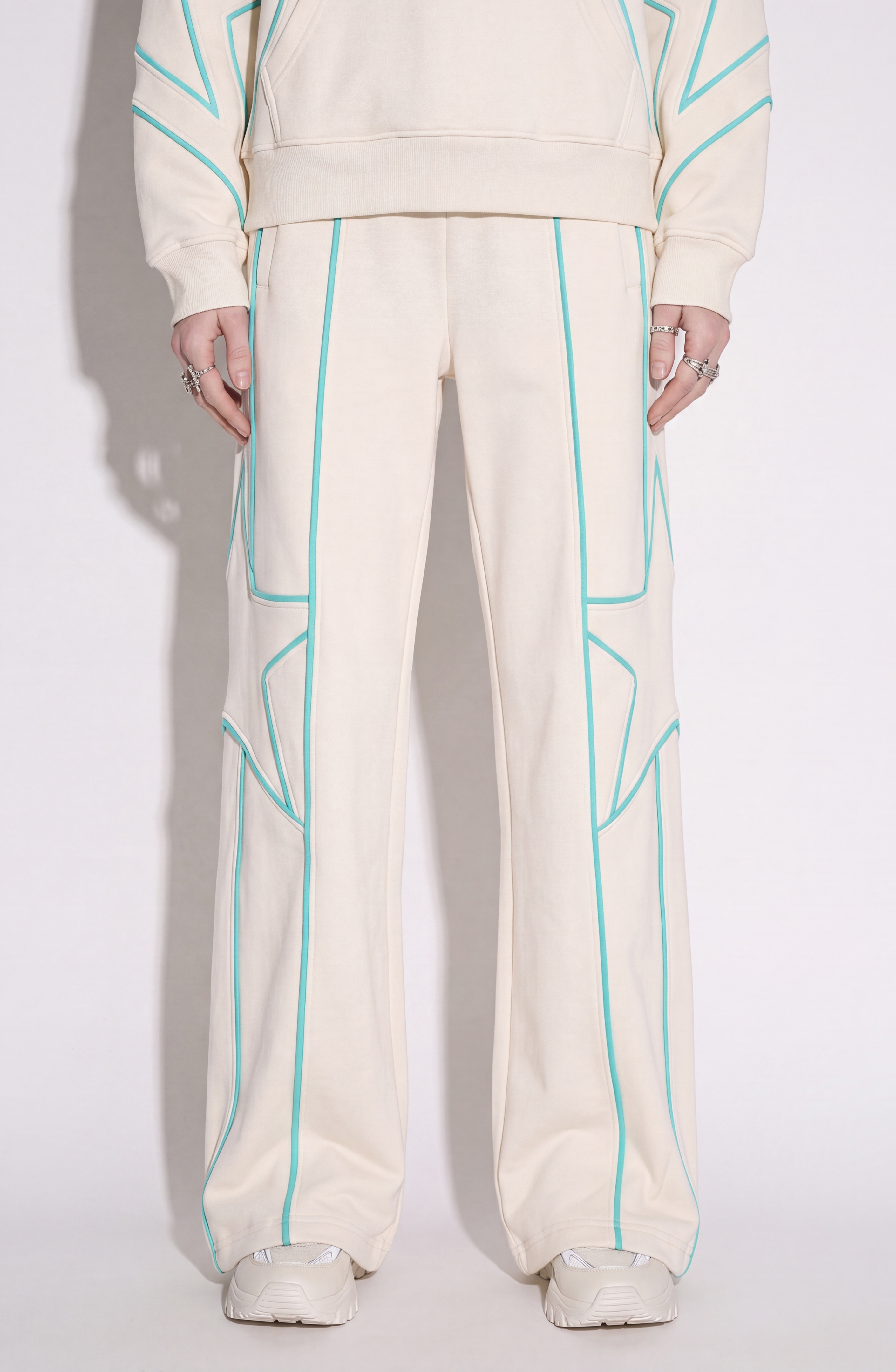 Styled in the LOULOU Embroidered Star Sweatpants—cream-colored wide-leg sweatpants crafted from soft cotton jersey with turquoise outlined geometric patterns and subtle turquoise star embroidery—this look is paired with a matching hoodie, white sneakers, and multiple rings, set against a plain light backdrop.
