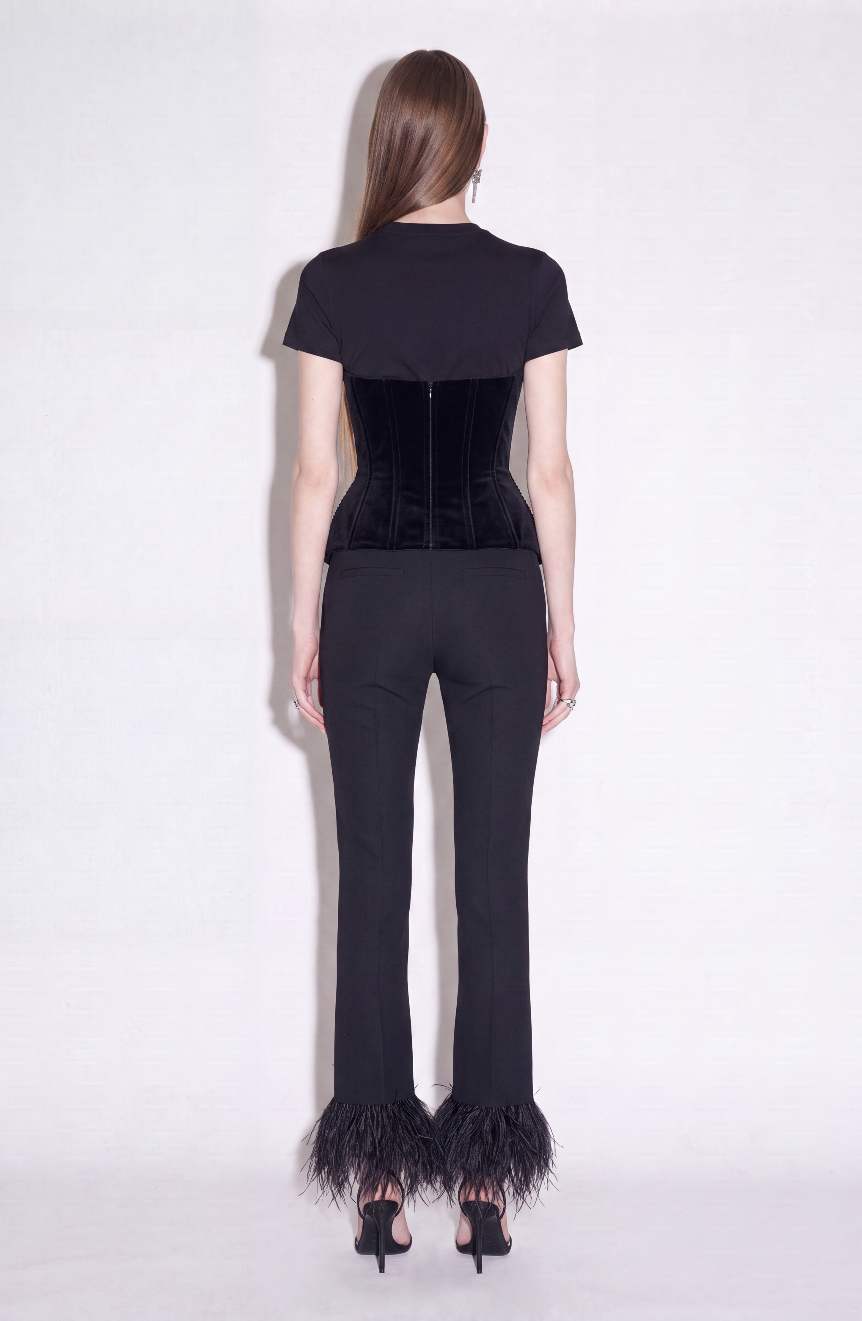 A woman stands with her back to the camera, wearing a fitted black t-shirt, a sleek black corset, and LOULOU’s Feather Trim Flared Trousers. The slim-fit trousers feature dramatic black feather trim and gently flare at the hem. She completes the look with black heels, while her straight, long brown hair cascades down her back. The background is plain white.