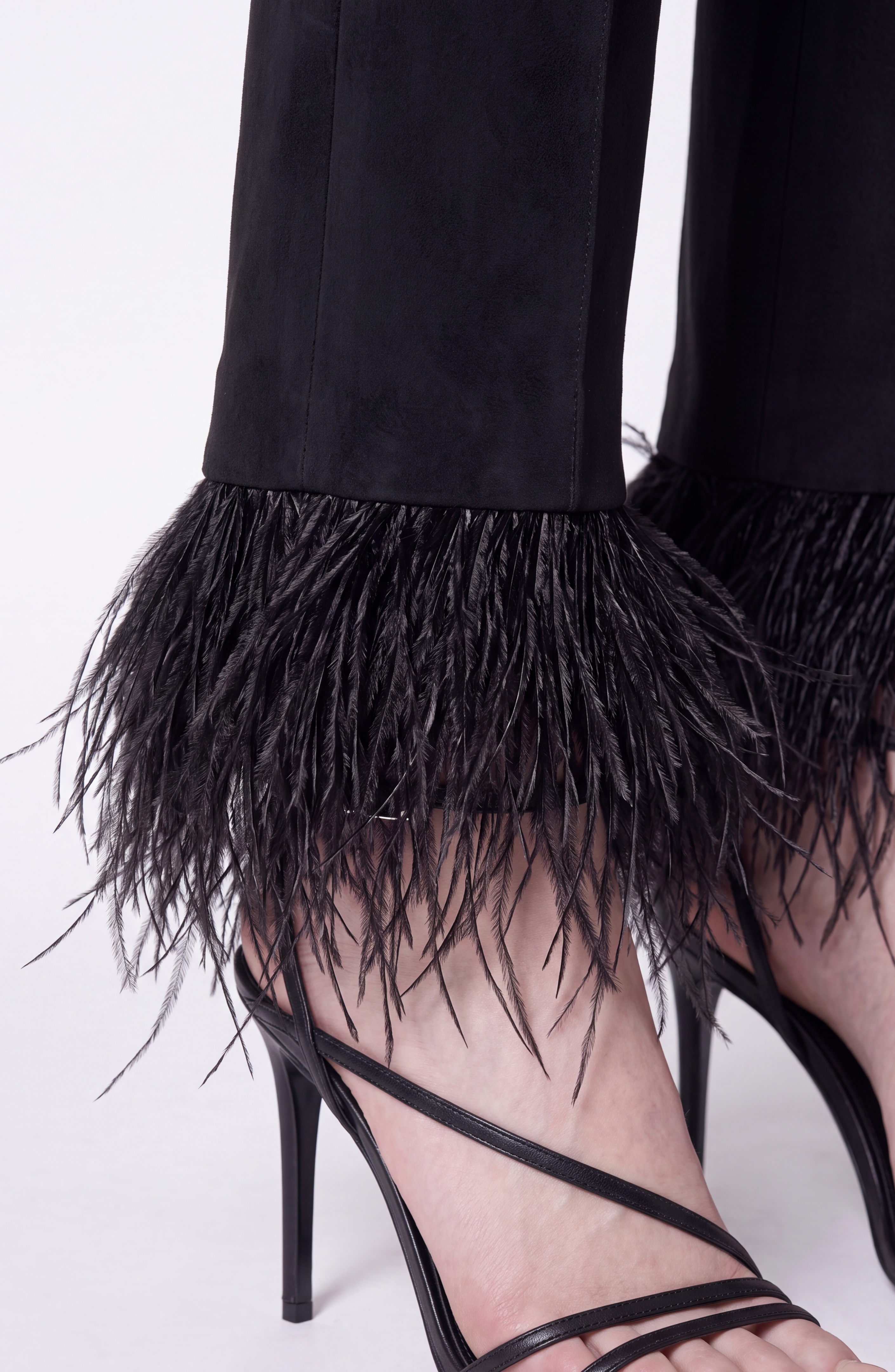 A close-up of the LOULOU Feather Trim Flared Trousers, featuring black feathered hems, styled with black strappy high-heeled sandals against a white background.