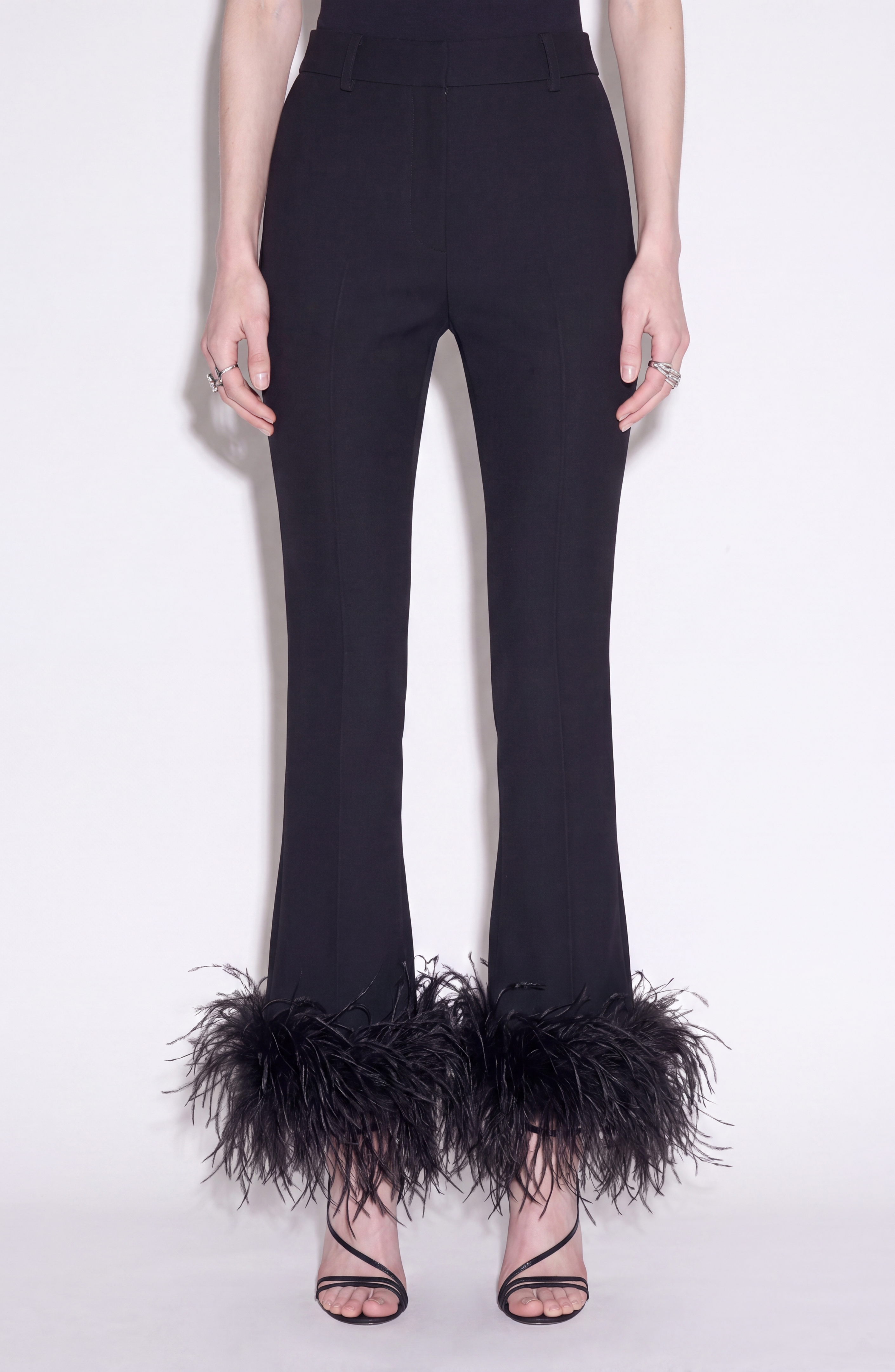 A person wearing LOULOU's Feather Trim Flared Trousers, featuring a high-waisted silhouette with black strappy high-heeled sandals, stands against a plain white background.