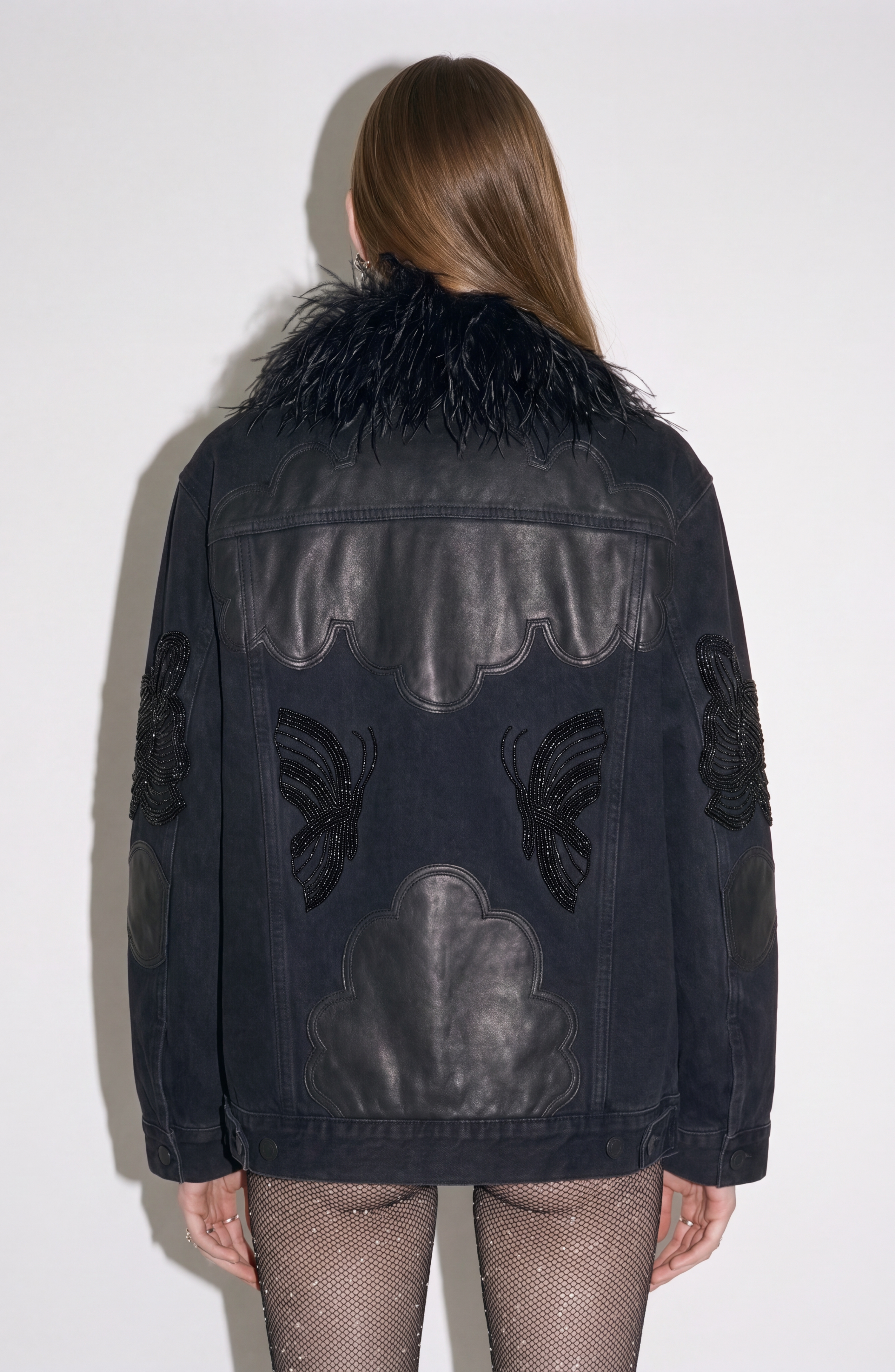 A person shown from behind wearing the LOULOU Feather-Trimmed Appliqué Denim Jacket, featuring an oversized fit, a feather collar, leather cloud-shaped appliqué patches, and beaded butterfly details on the back and sleeves.