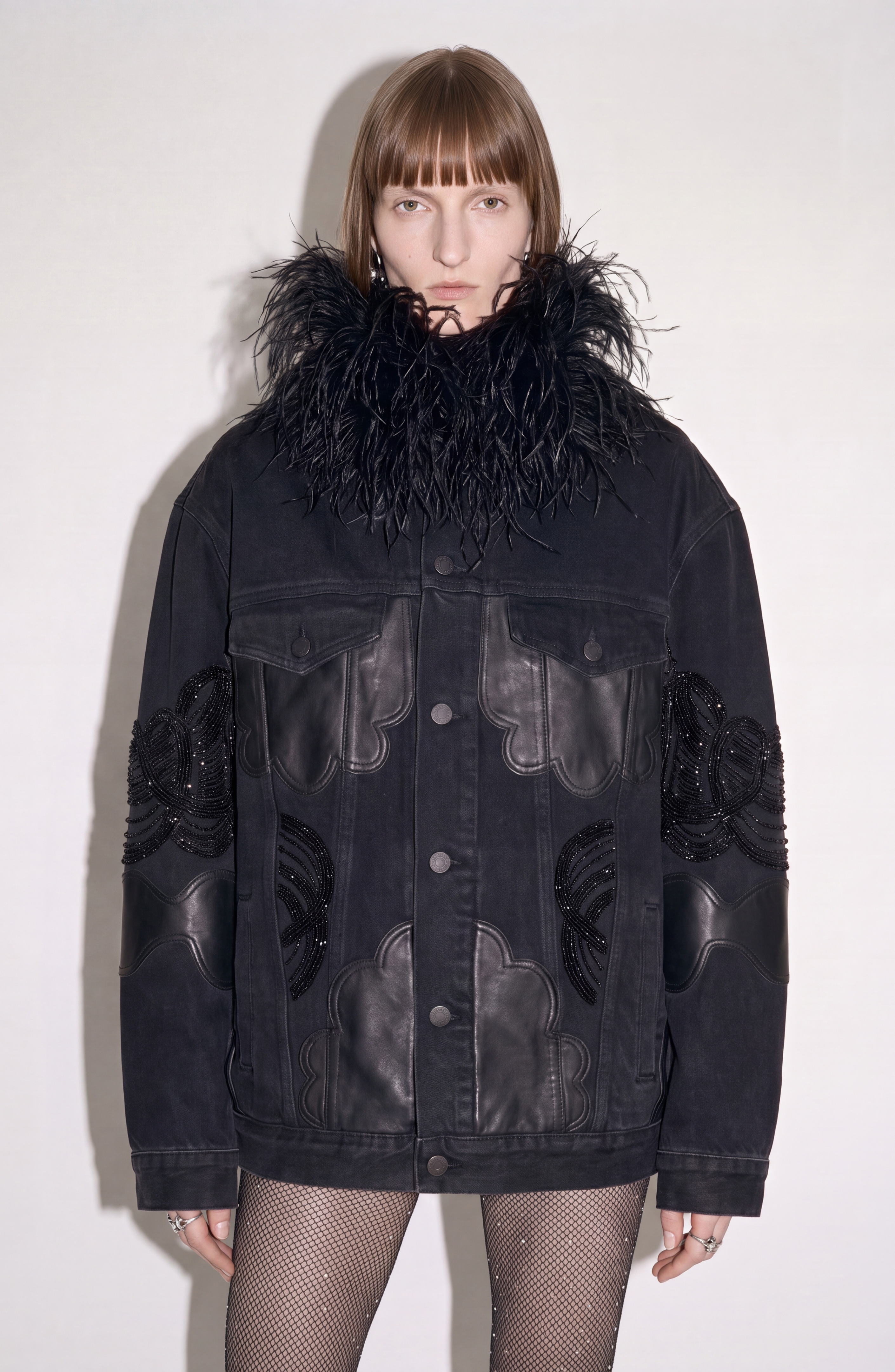 A person wearing the LOULOU Feather-Trimmed Appliqué Denim Jacket—an oversized black piece with a dramatic feather collar, intricate leather and beaded details—paired with fishnet stockings, stands against a plain light background, looking directly at the camera.