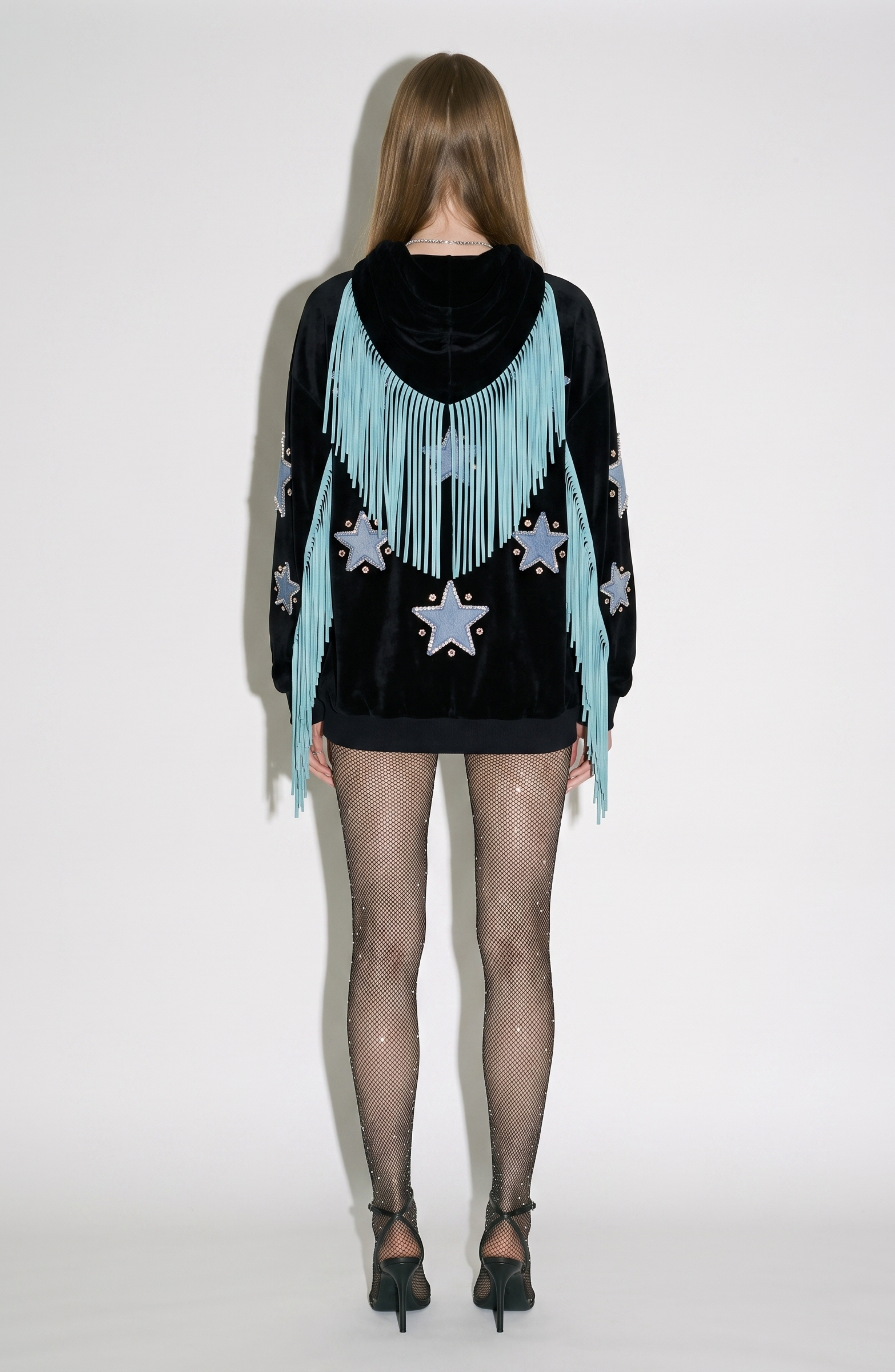 A person stands facing away against a plain white background, wearing the LOULOU Fringed Star Velvet Hoodie—black velvet with blue fringe and star patches—styled with sheer polka-dot tights and black heels.