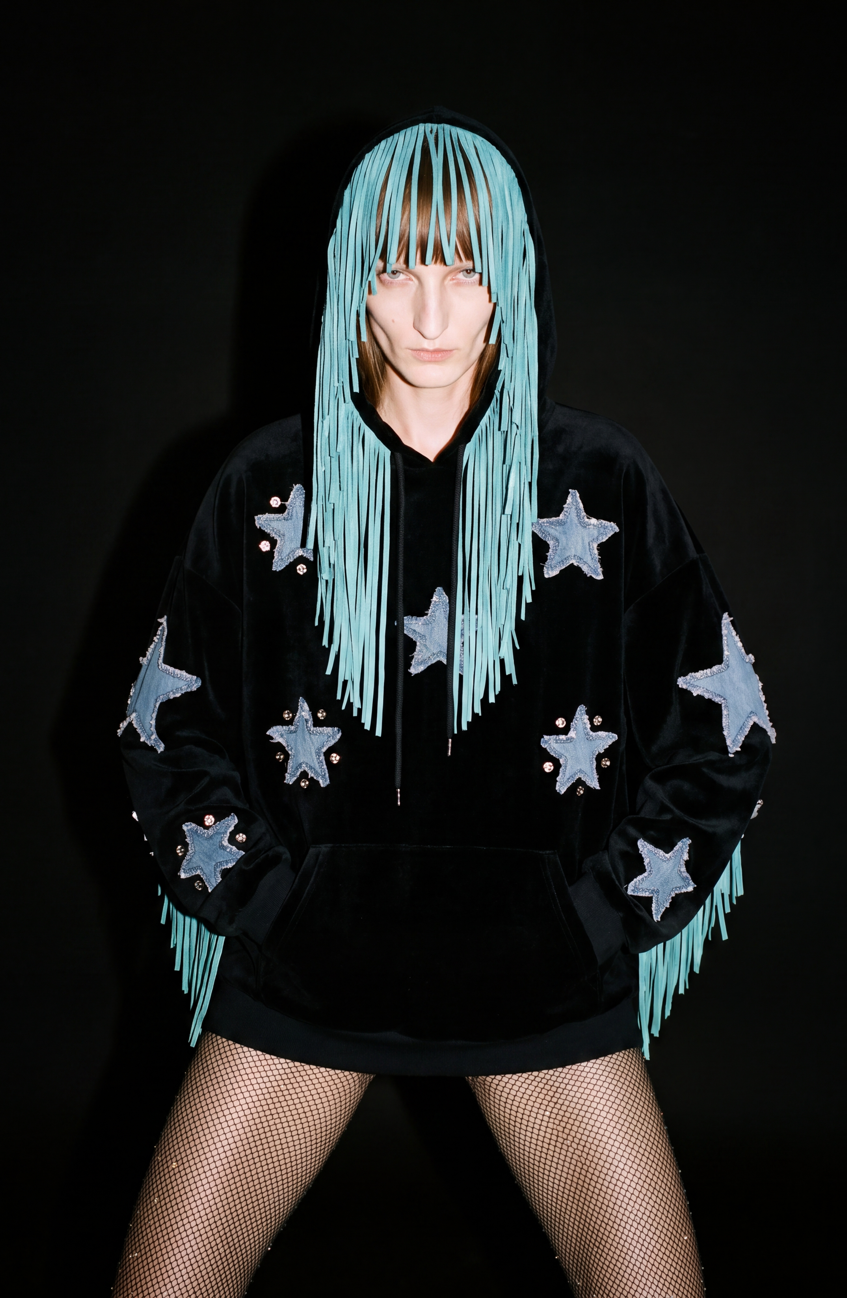 Wearing the LOULOU Fringed Star Velvet Hoodie—featuring denim star appliqués and cascading long blue fringe—a model stands against a black background with hands in pockets, paired with fishnet tights.