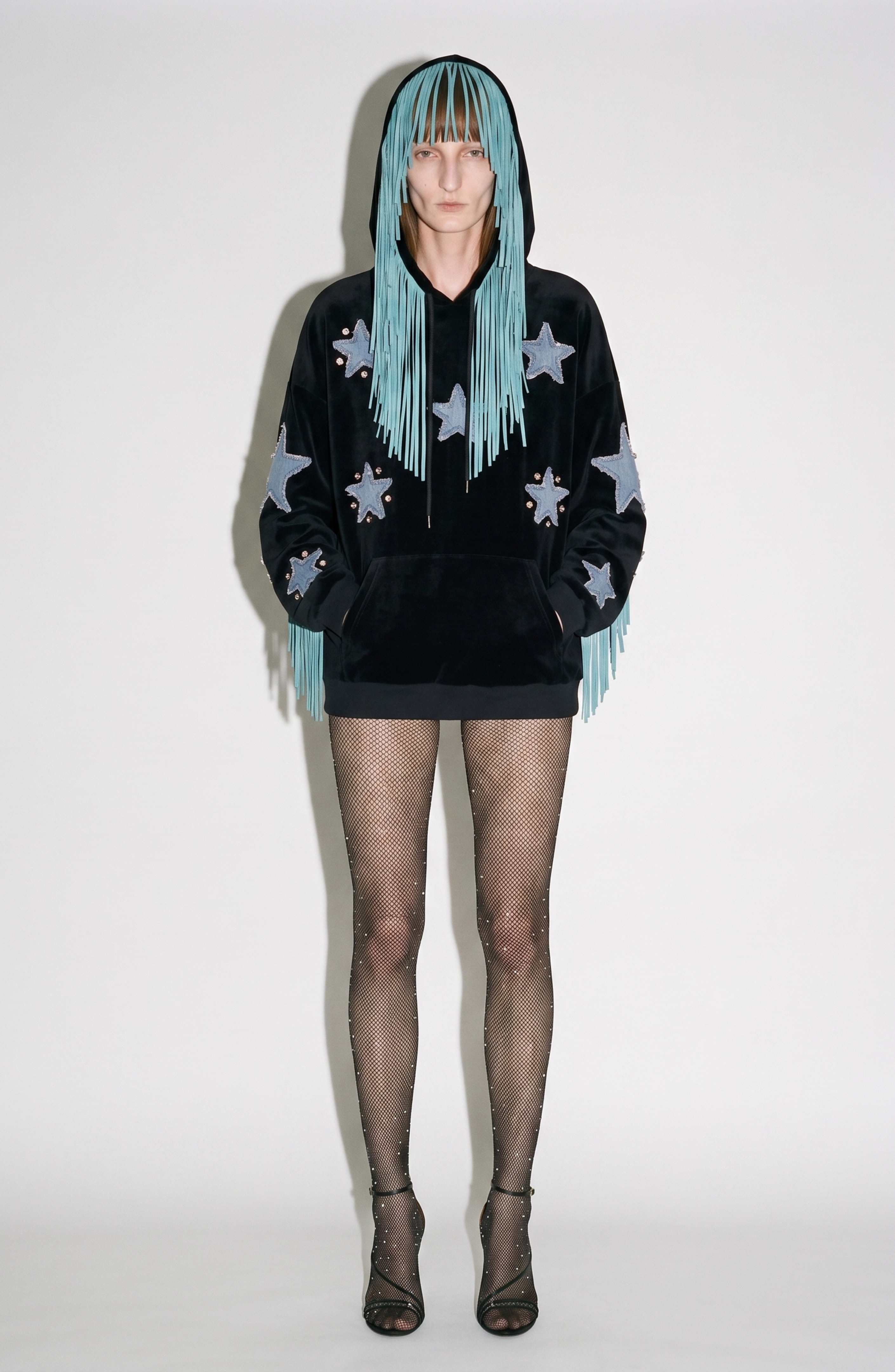 A model wears the LOULOU Fringed Star Velvet Hoodie—crafted from black velvet and detailed with star patches and long teal fringes that cascade over the hood and face—paired with black fishnet stockings embellished with crystals and black high-heeled sandals, standing against a plain background.