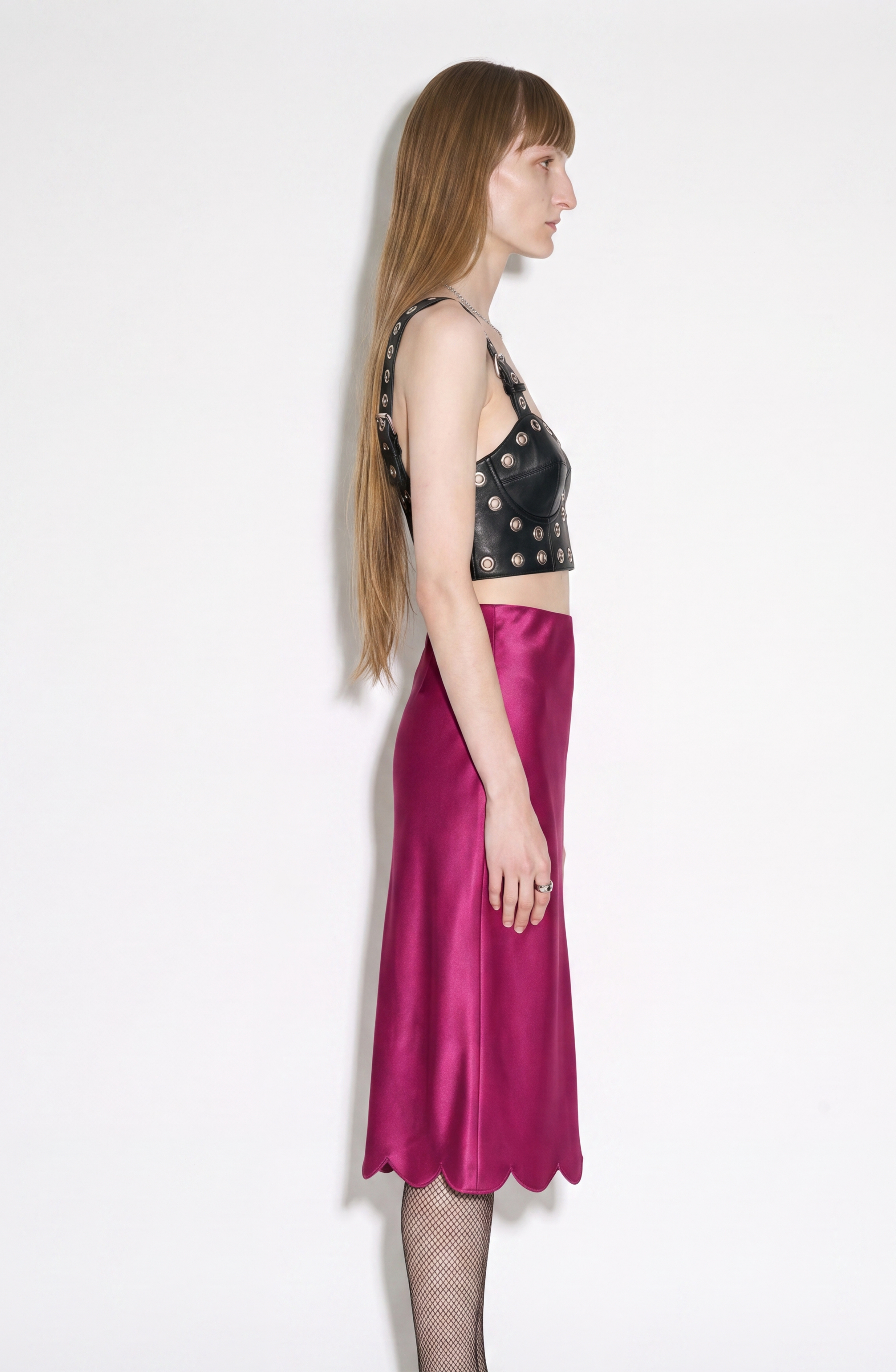A person with long straight hair stands in profile against a white background, wearing a sleeveless black studded leather top, LOULOU's Fuchsia Silk Skirt with a scalloped hemline, and fishnet stockings.