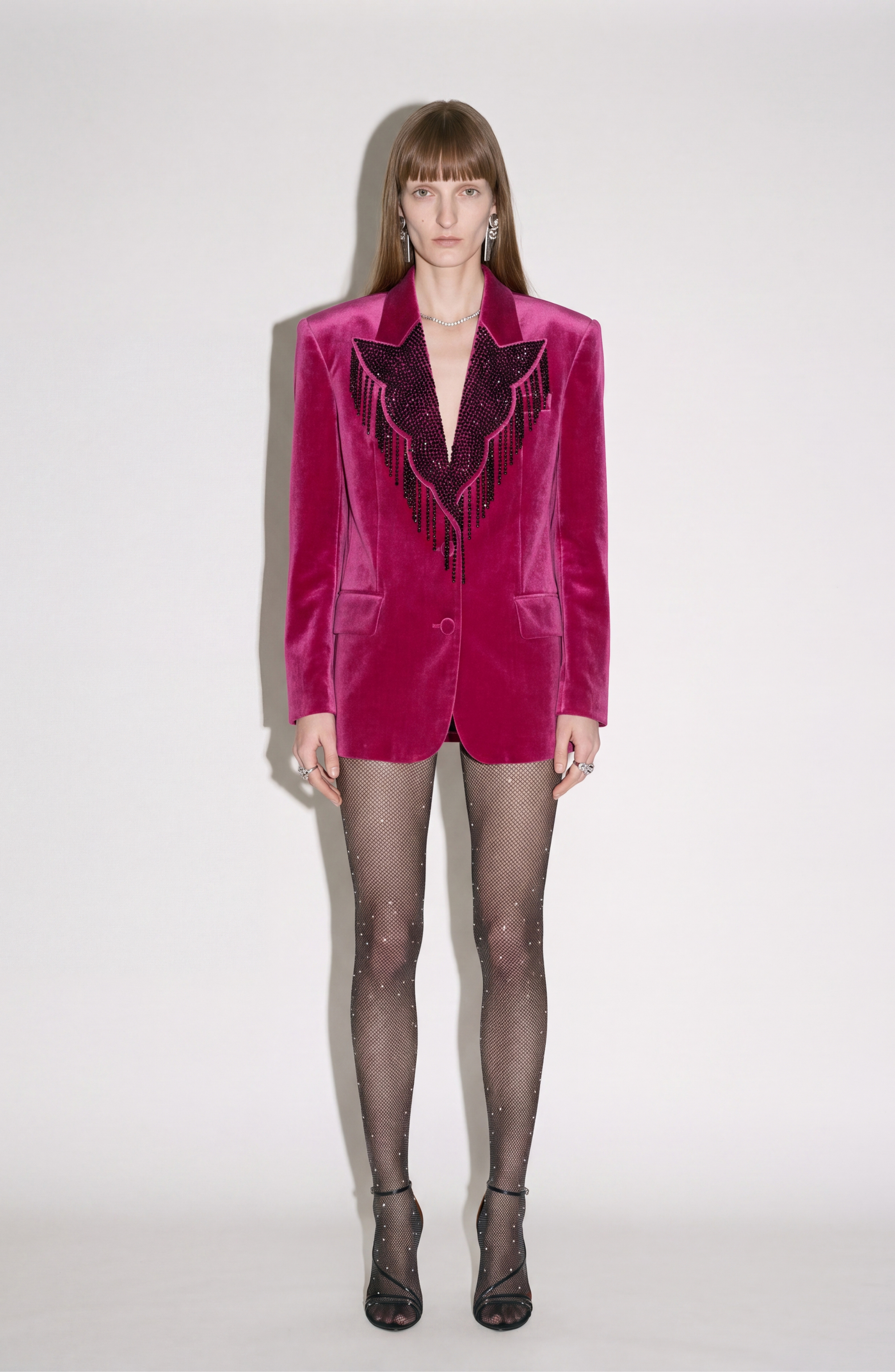 A person stands against a plain white background wearing the LOULOU Fuchsia Velvet Blazer with sharp shoulders and black beaded fringe, sheer sparkly black tights, and black open-toe heels.