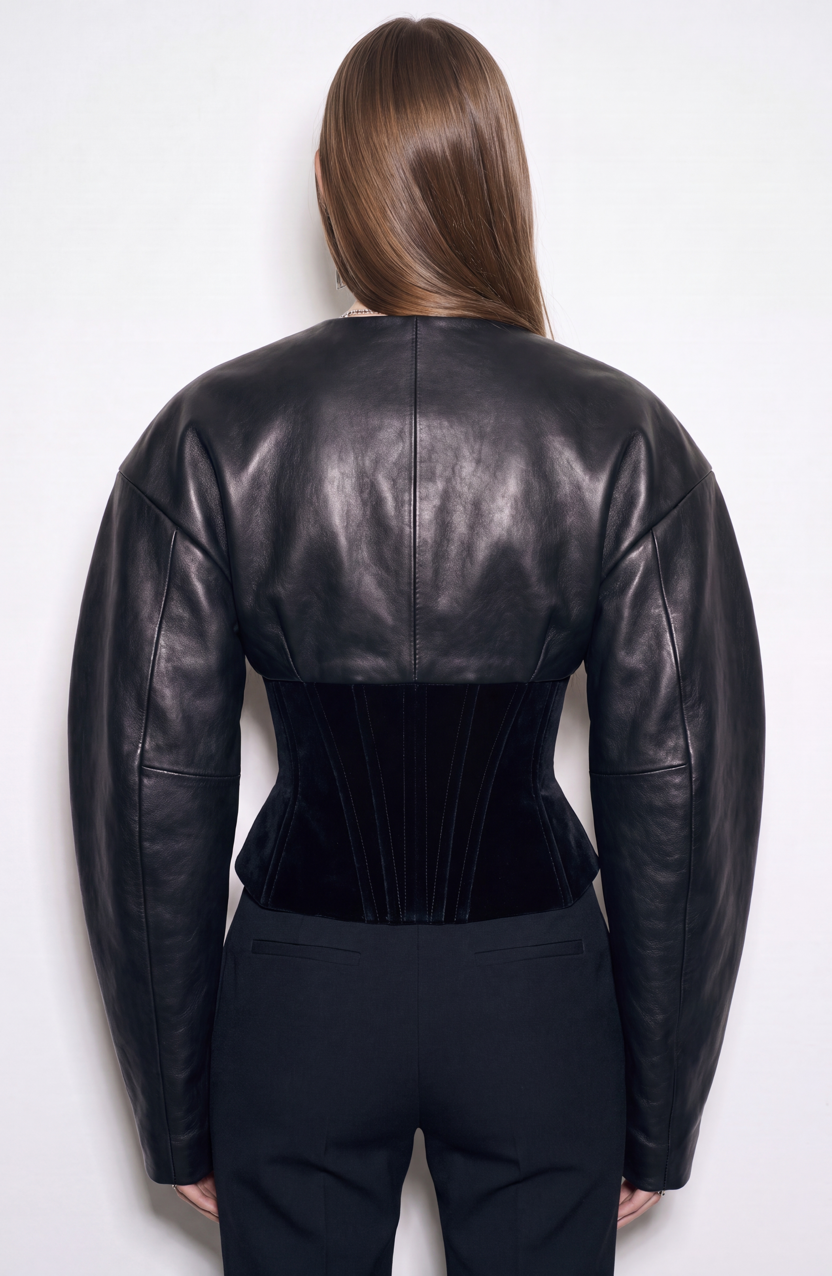 A person with straight brown hair wears the LOULOU Leather And Velvet Corset Jacket—a structured black leather piece with exaggerated shoulders, a fitted waist, and subtly voluminous sleeves—paired with black pants while standing against a plain white background.