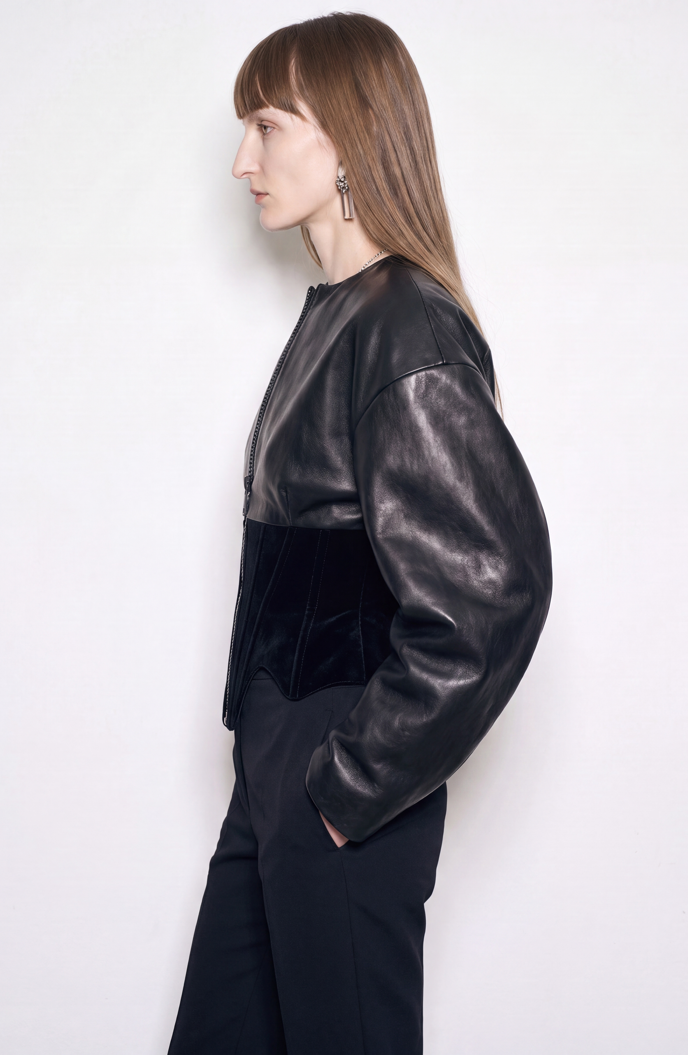 A woman with straight brown hair stands in profile against a plain white background, wearing the LOULOU Leather and Velvet Corset Jacket featuring voluminous sleeves, paired with black pants. She accessorizes with long earrings and has a neutral expression.