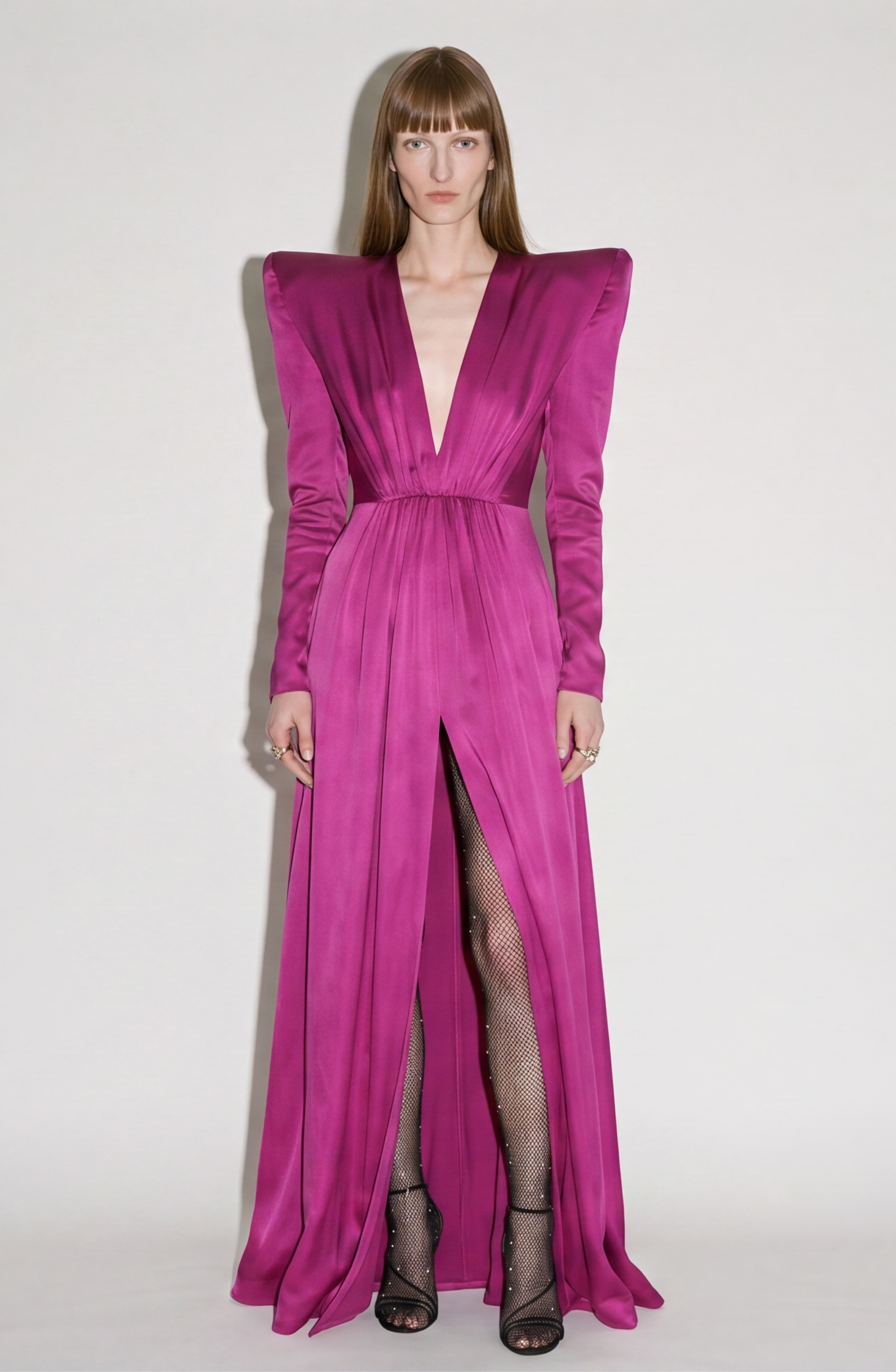 A model showcases the LOULOU Magenta Satin Gown featuring structured shoulders, a deep V neckline, and a high front slit, styled with black fishnet stockings and open-toe heels against a plain light background.