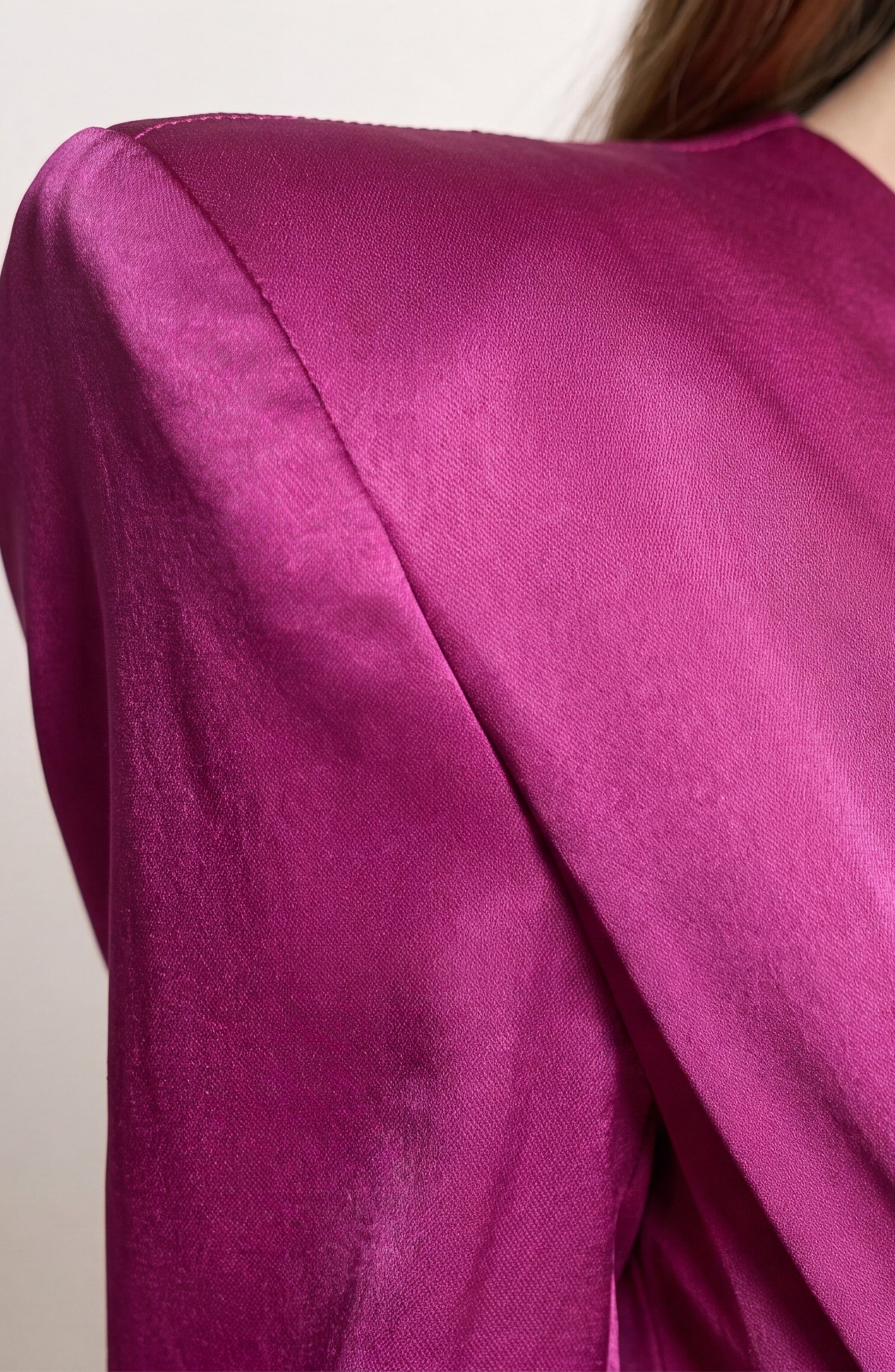 Close-up of a person in the LOULOU Magenta Satin Gown, featuring structured padded shoulders and a deep V neckline that highlight the smooth fabric and bold, full-length silhouette.