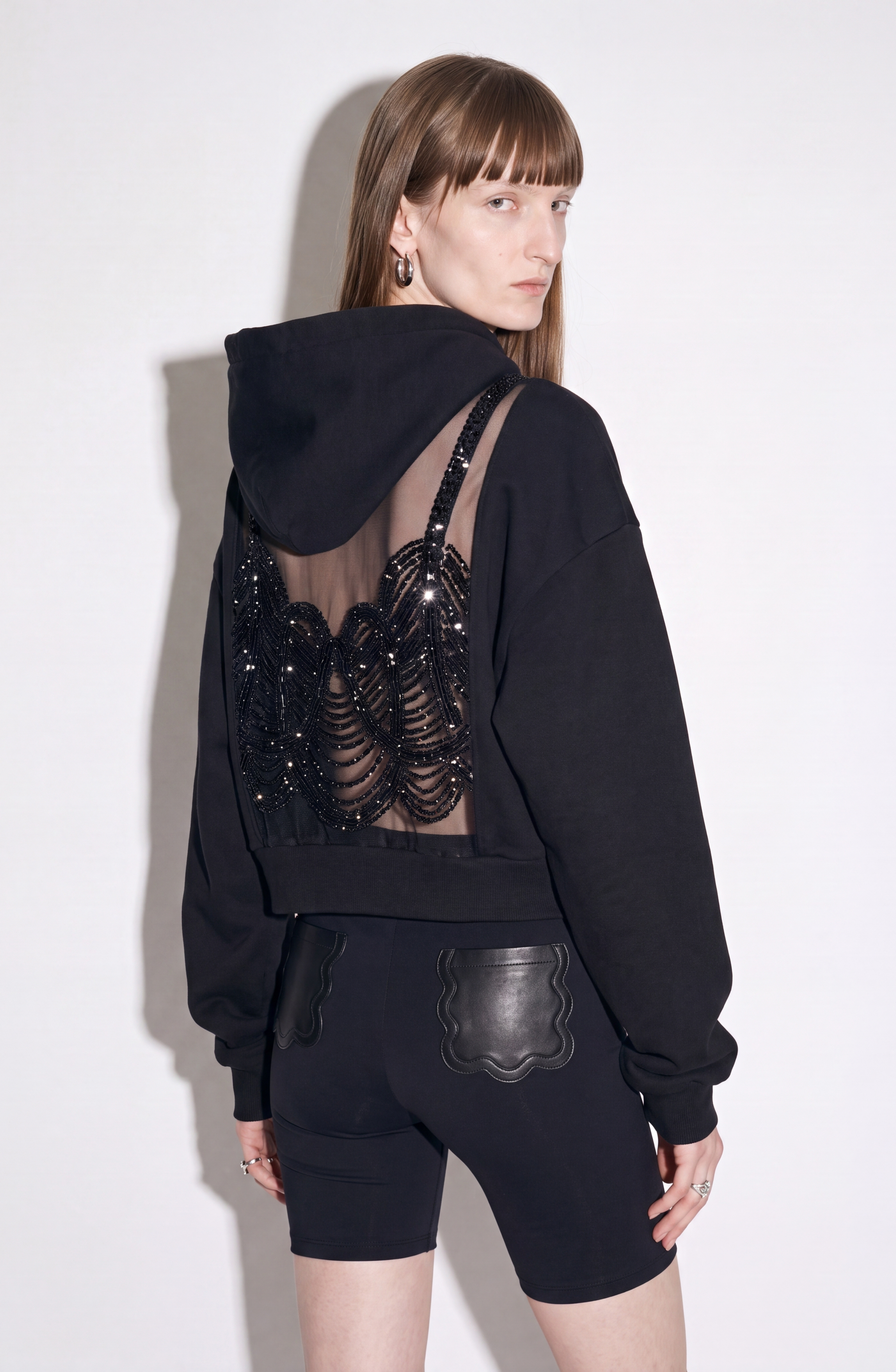 A model stands against a plain white background, facing backward and looking over their shoulder, wearing LOULOU’s Scalloped Leather Pocket Shorts—high-waisted black shorts with scalloped leather patch pockets—paired with a black hoodie featuring a sheer, sequined back panel.