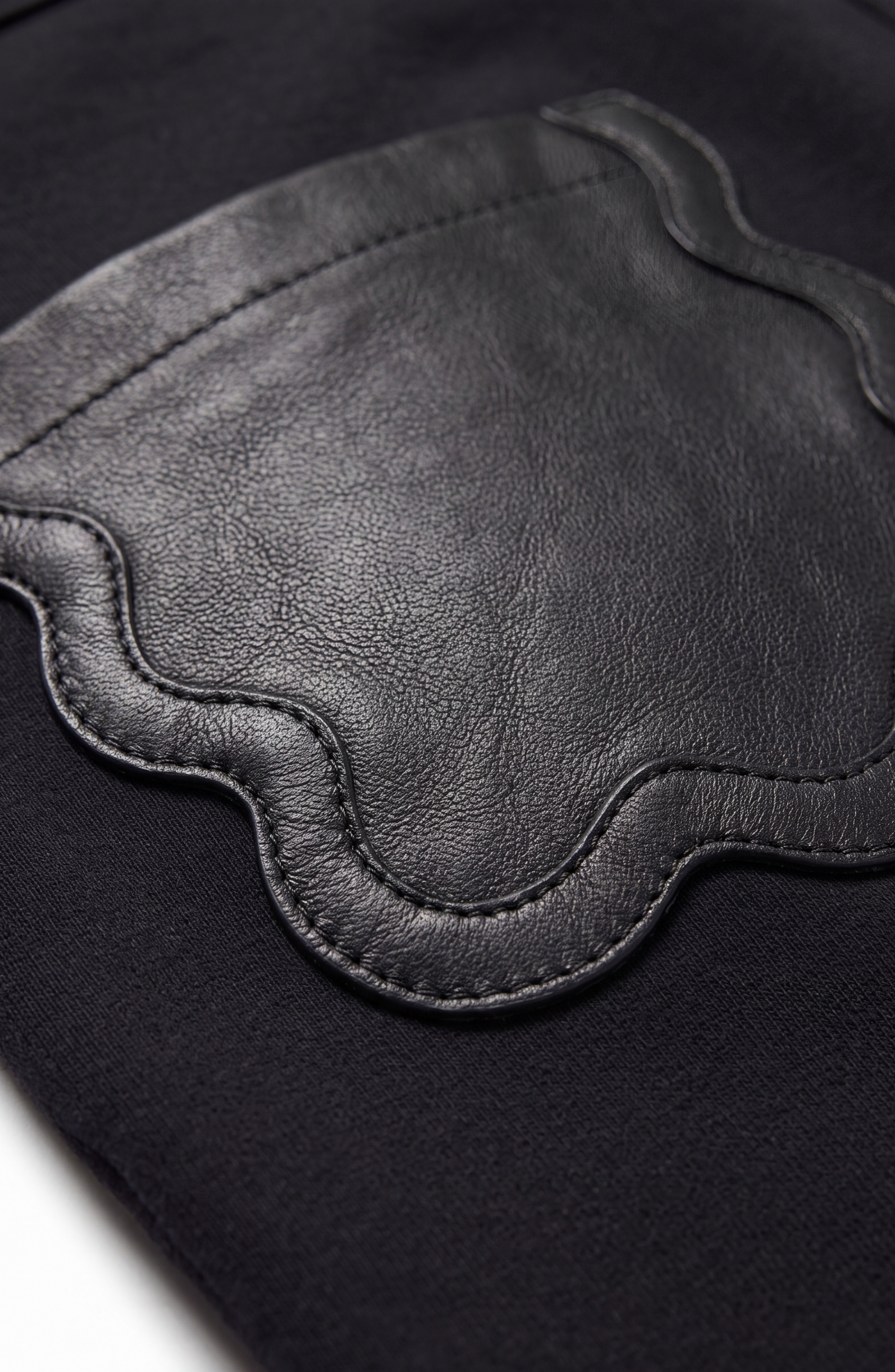 Close-up of a black scalloped leather patch with a wavy edge, sewn onto a dark stretch jersey fabric background. The patch features a smooth, textured surface and is outlined with visible stitching, echoing the signature pocket design of LOULOU’s Scalloped Leather Pocket Shorts.