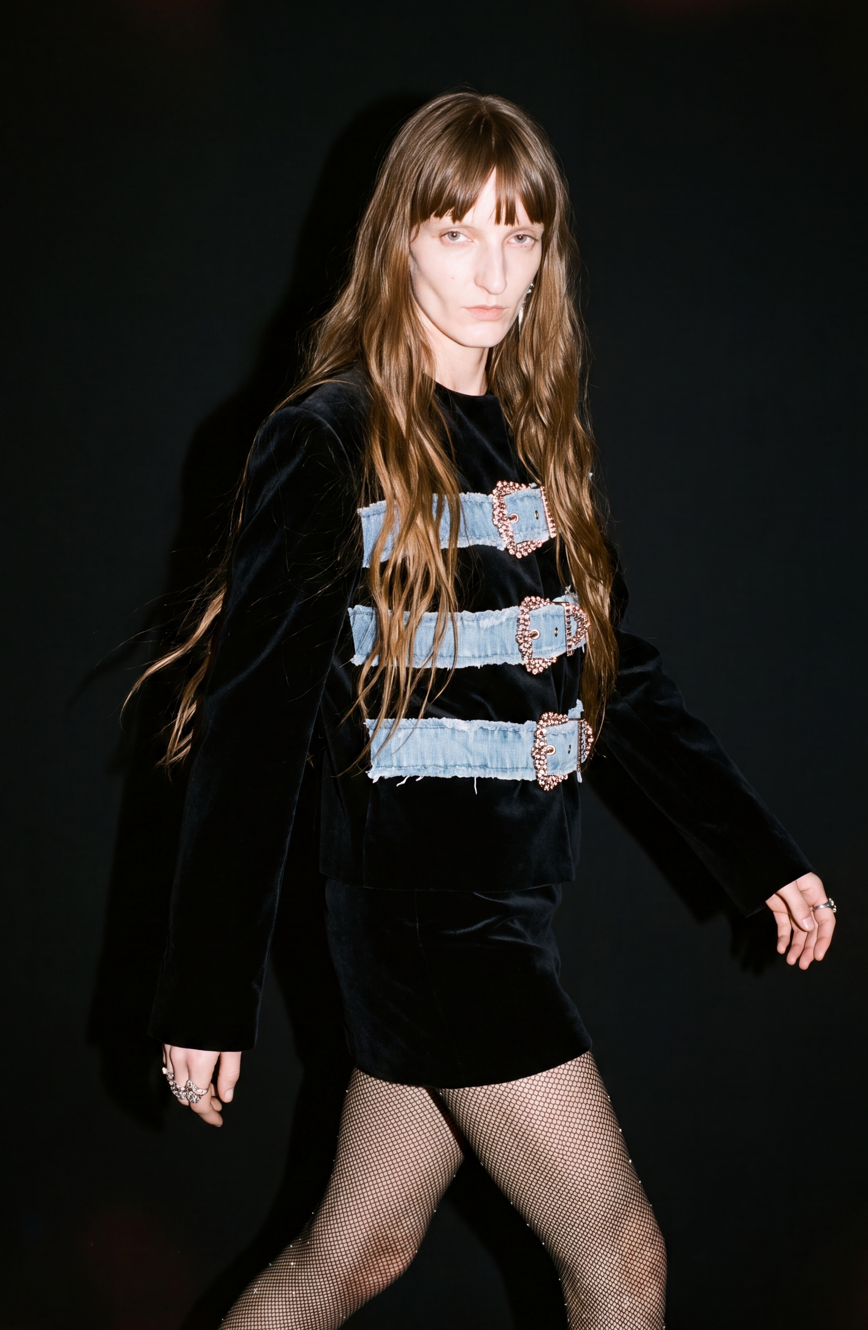 A person with long brown hair walks in front of a black background, wearing the LOULOU Sculpted Black Velvet Mini Skirt styled with three large jeweled belt-buckles and fishnet stockings.