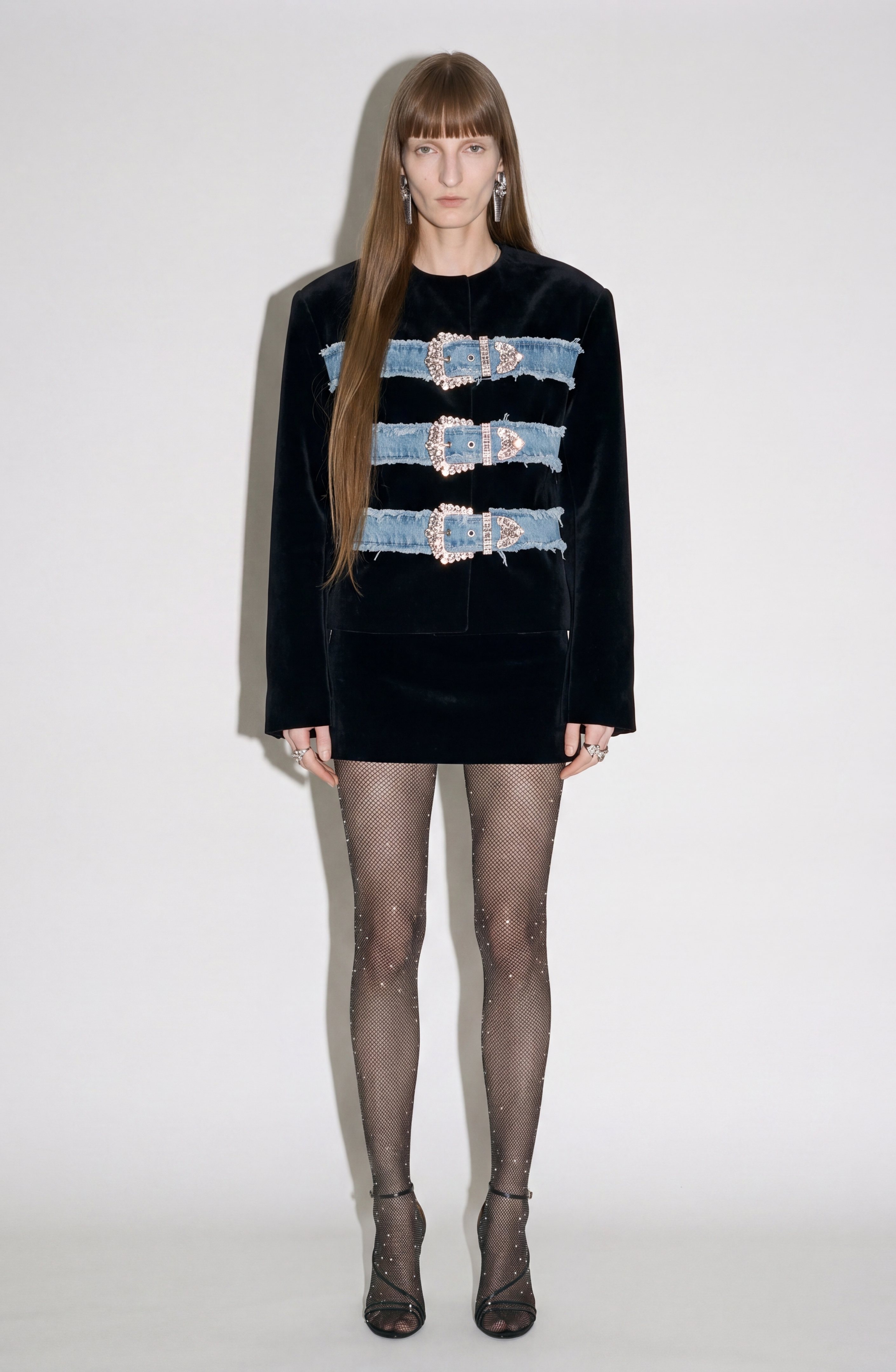 A person stands against a plain background wearing the LOULOU Sculpted Black Velvet Mini Skirt, styled with three denim straps across the chest, sheer black rhinestone tights, and black open-toe heels.