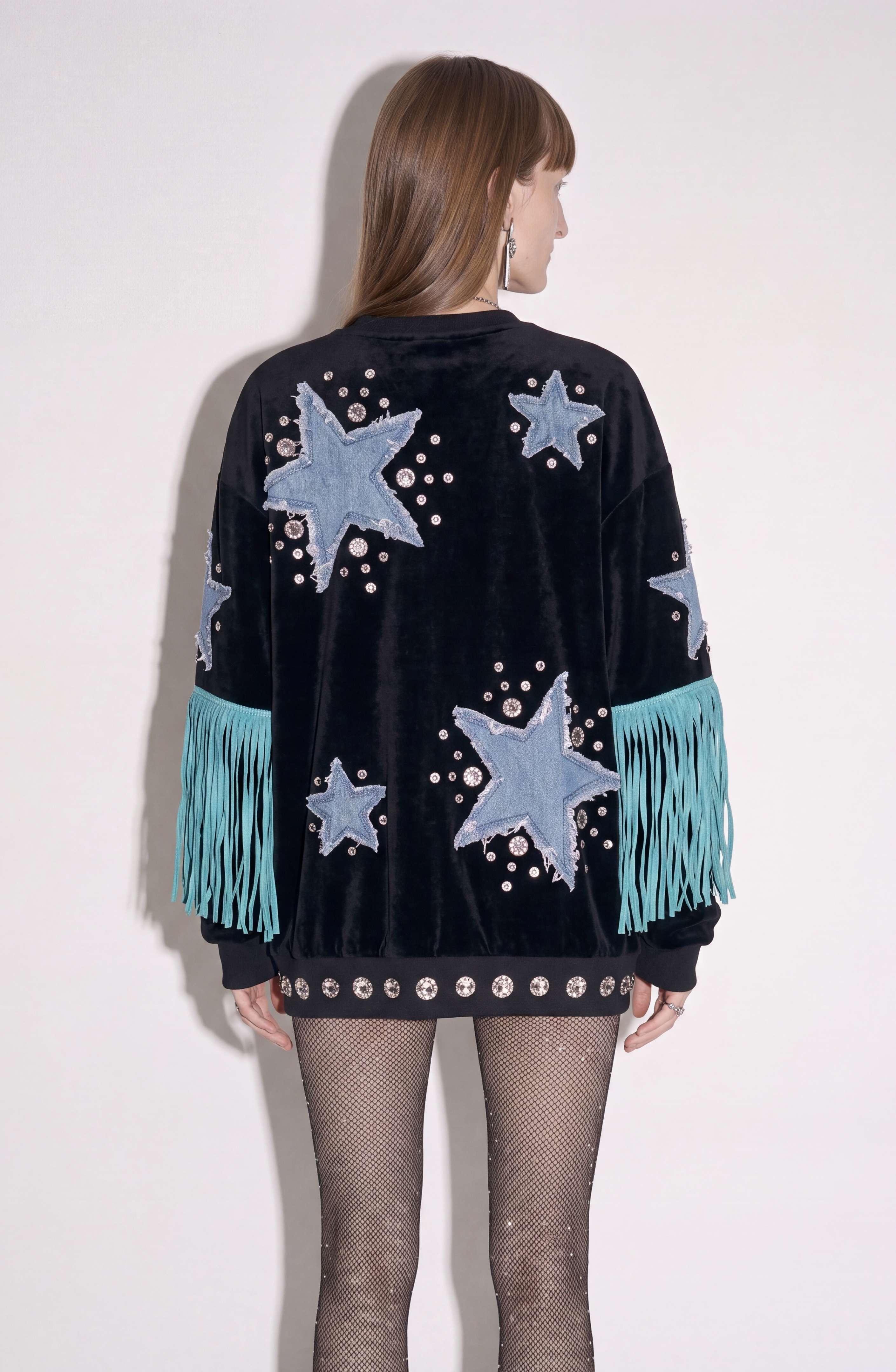 A person stands facing away, wearing the LOULOU Star Appliqué Velvet Sweatshirt—black with blue denim star appliqués, silver studs, and turquoise fringe on the sleeves—paired with sparkly fishnet tights.