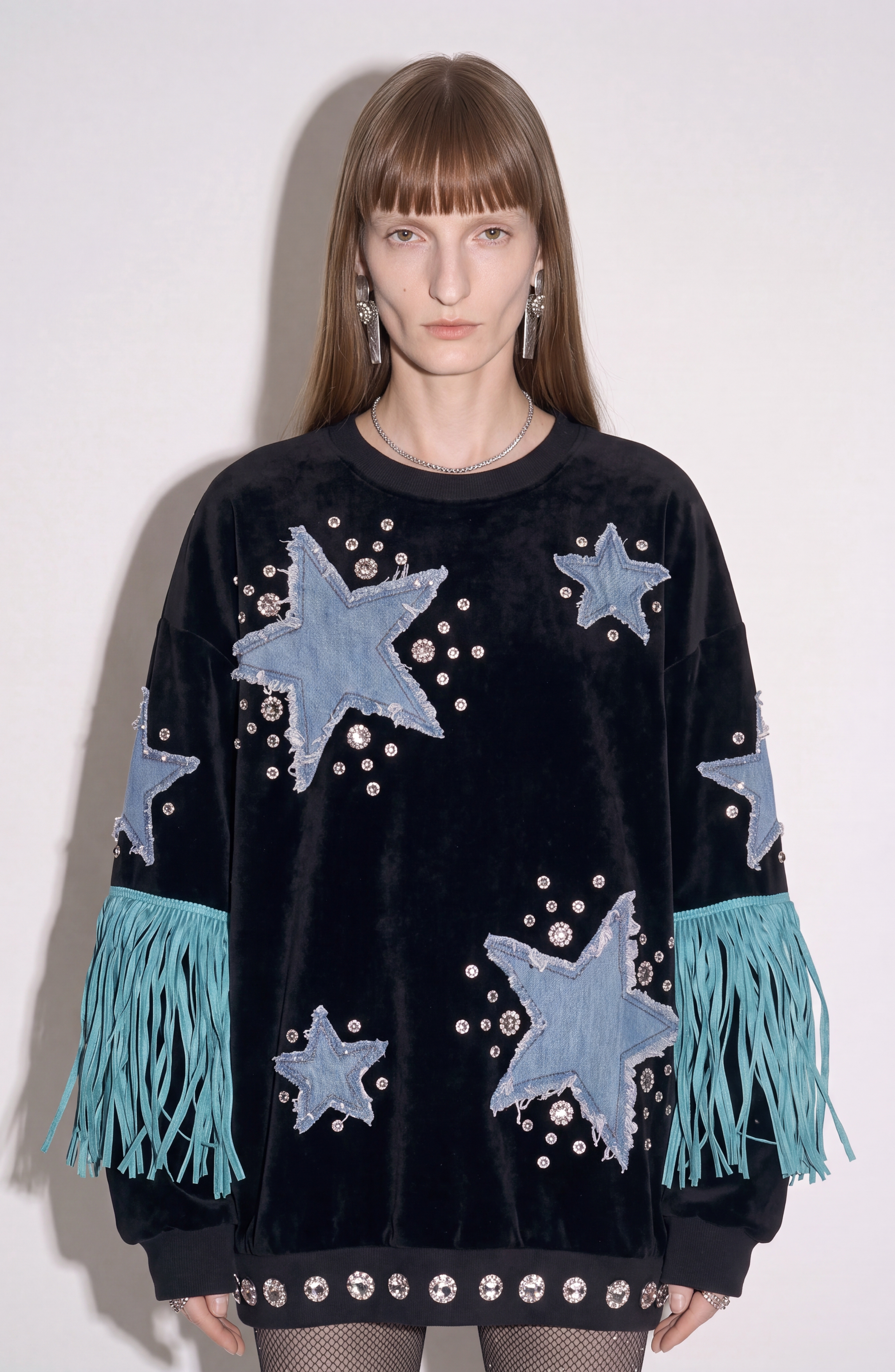A model stands against a plain white background wearing the LOULOU Star Appliqué Velvet Sweatshirt, featuring black velvet fabric embellished with blue denim star appliqués, silver studs, and turquoise fringe on the sleeves.