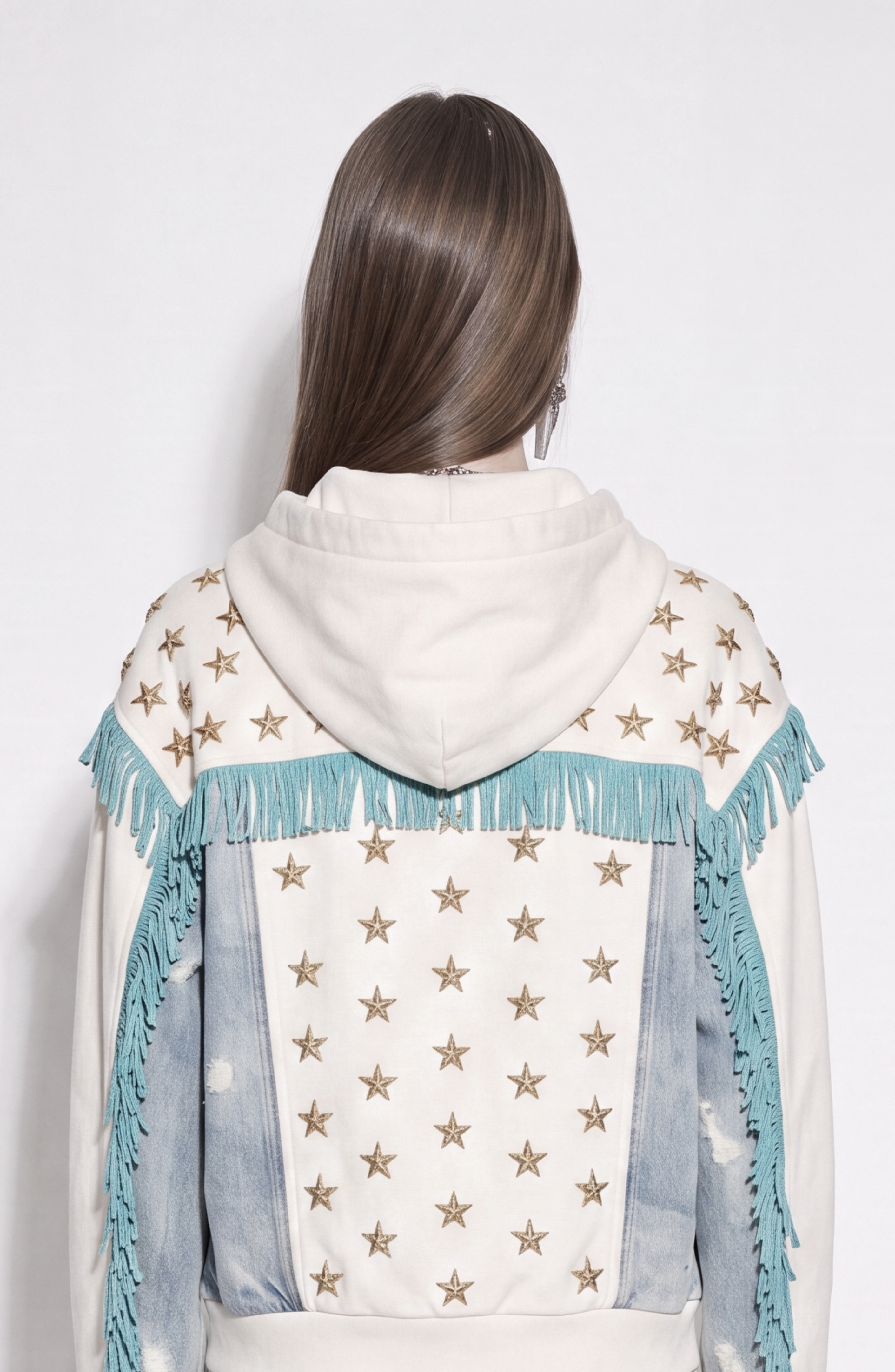 A person with long brown hair faces away, wearing the LOULOU Star Studded Fringed Zip Hoodie—a relaxed-fit, off-white cotton jersey zip hoodie detailed with gold stars and turquoise fringe on the shoulders. Light blue distressed denim panels accent the back and sleeves for a unique finish.