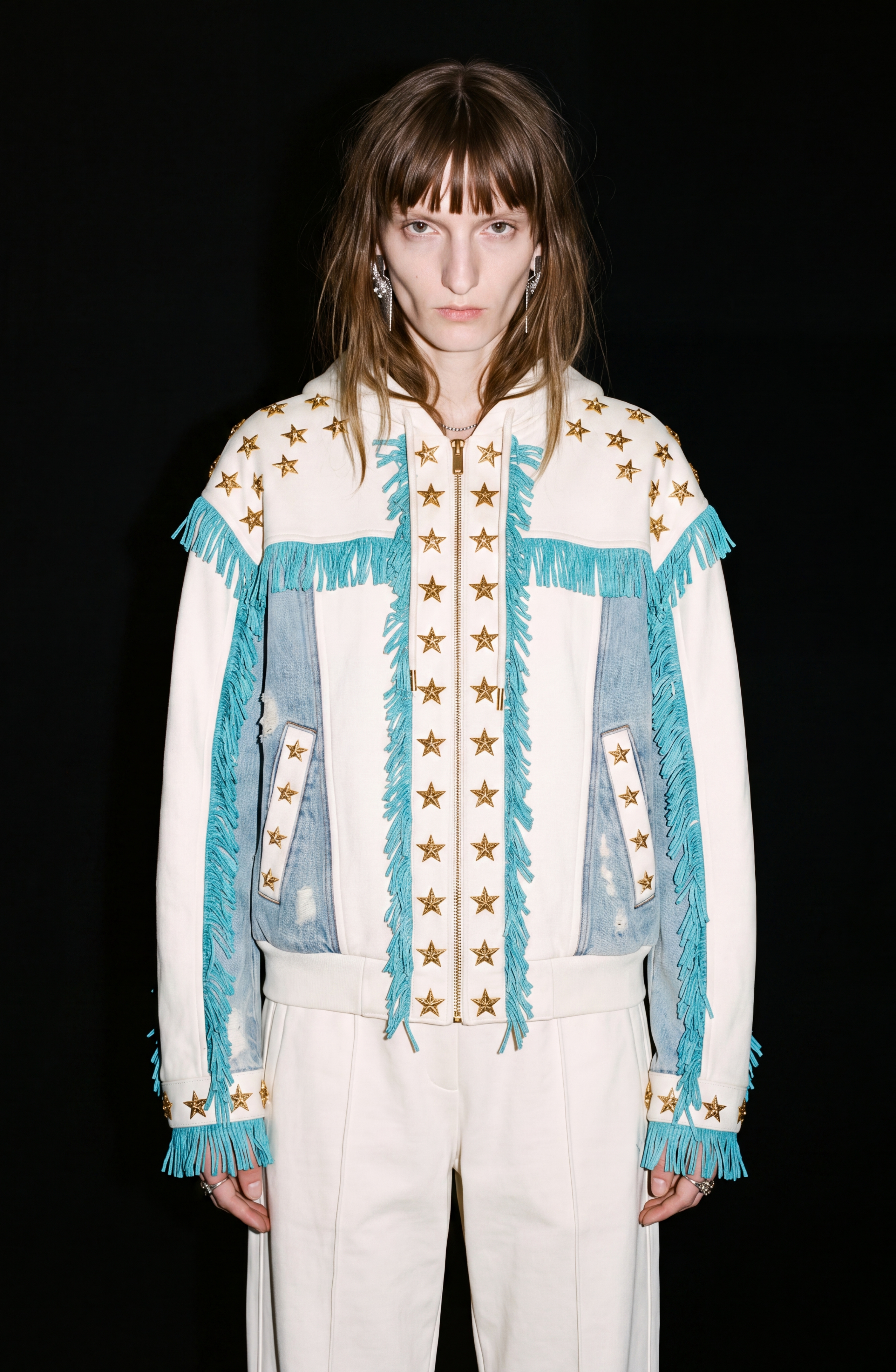 A person stands against a black background wearing the LOULOU Star Studded Fringed Zip Hoodie, featuring white fabric adorned with gold star studs and turquoise fringe. The contemporary look is completed with matching white pants, relaxed-fit zip hoodie details, and distressed denim panels for an edgy finish.