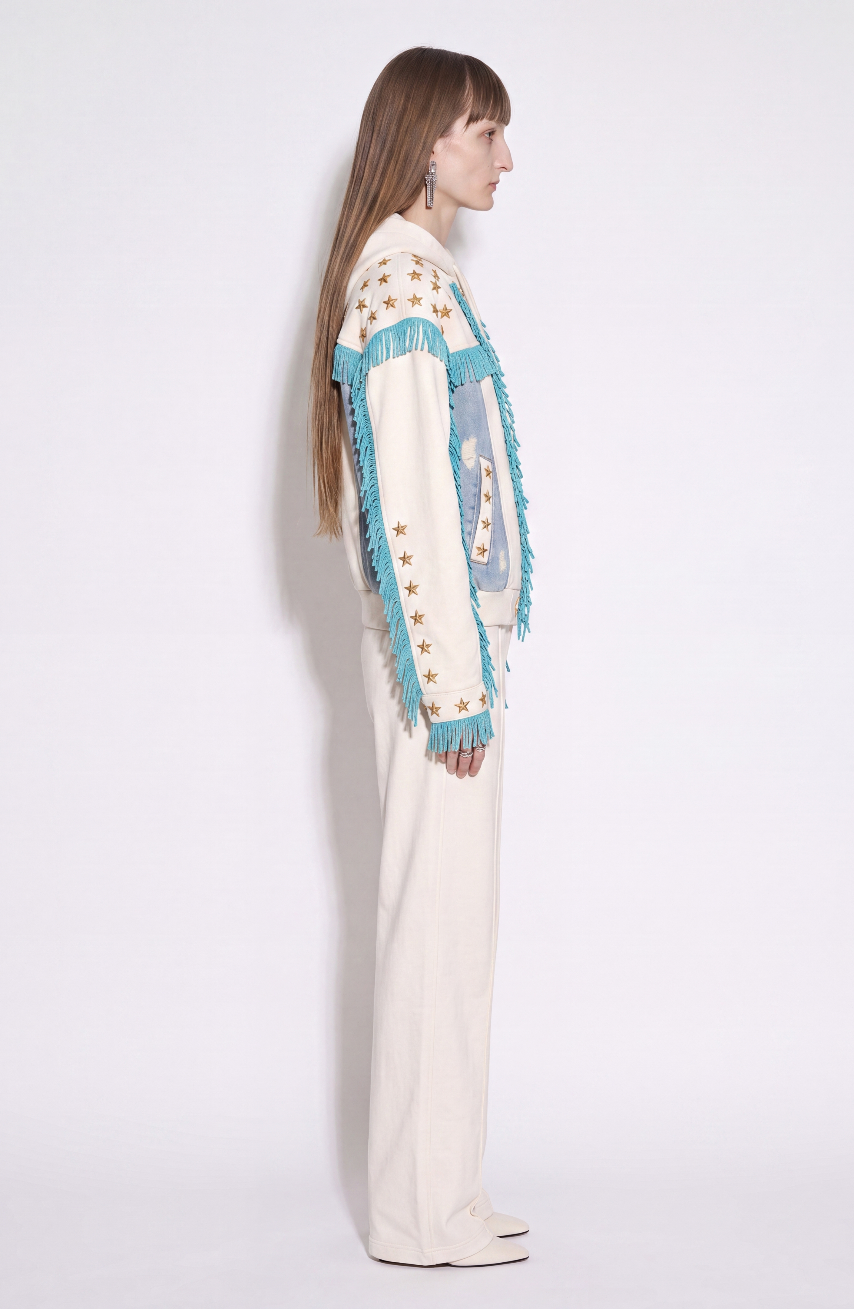 A person stands in profile against a plain white background, wearing the LOULOU Star Studded Fringed Zip Hoodie—a cream piece featuring blue fringe and star details with distressed denim panels. Their long brown hair complements the look, along with dangling earrings.