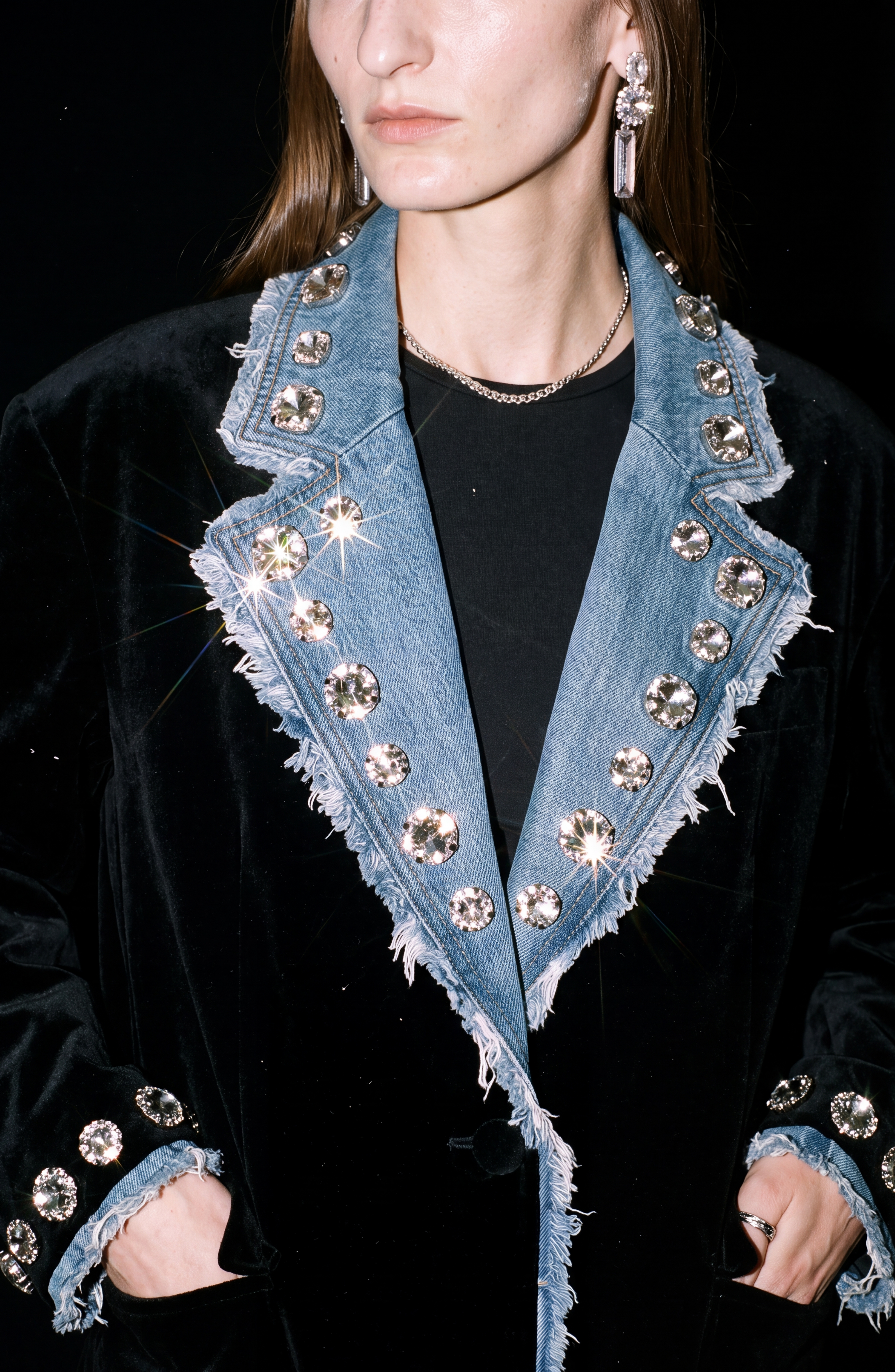 A person wearing the LOULOU Velvet And Denim Crystal Blazer—crafted from black velvet with frayed denim lapels and cuffs, adorned with large sparkling glass crystal embellishments—styled with a black shirt, shiny earrings, and a chain necklace.