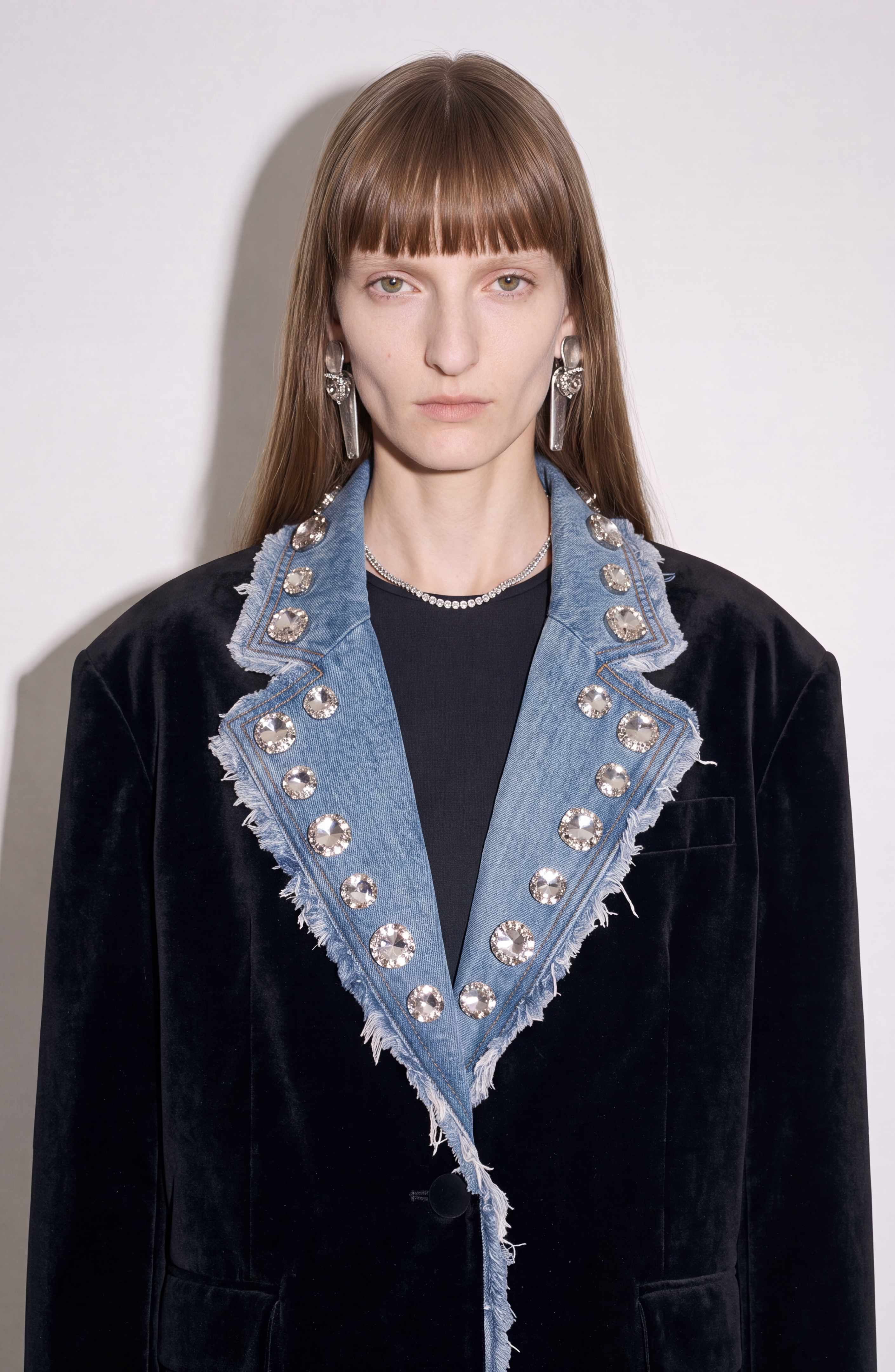 A model with straight brown hair and bangs showcases the Velvet And Denim Crystal Blazer by LOULOU, a black velvet piece distinguished by its frayed denim lapel embellished with dazzling glass crystals. She completes her look with long metallic drop earrings and a delicate crystal necklace.