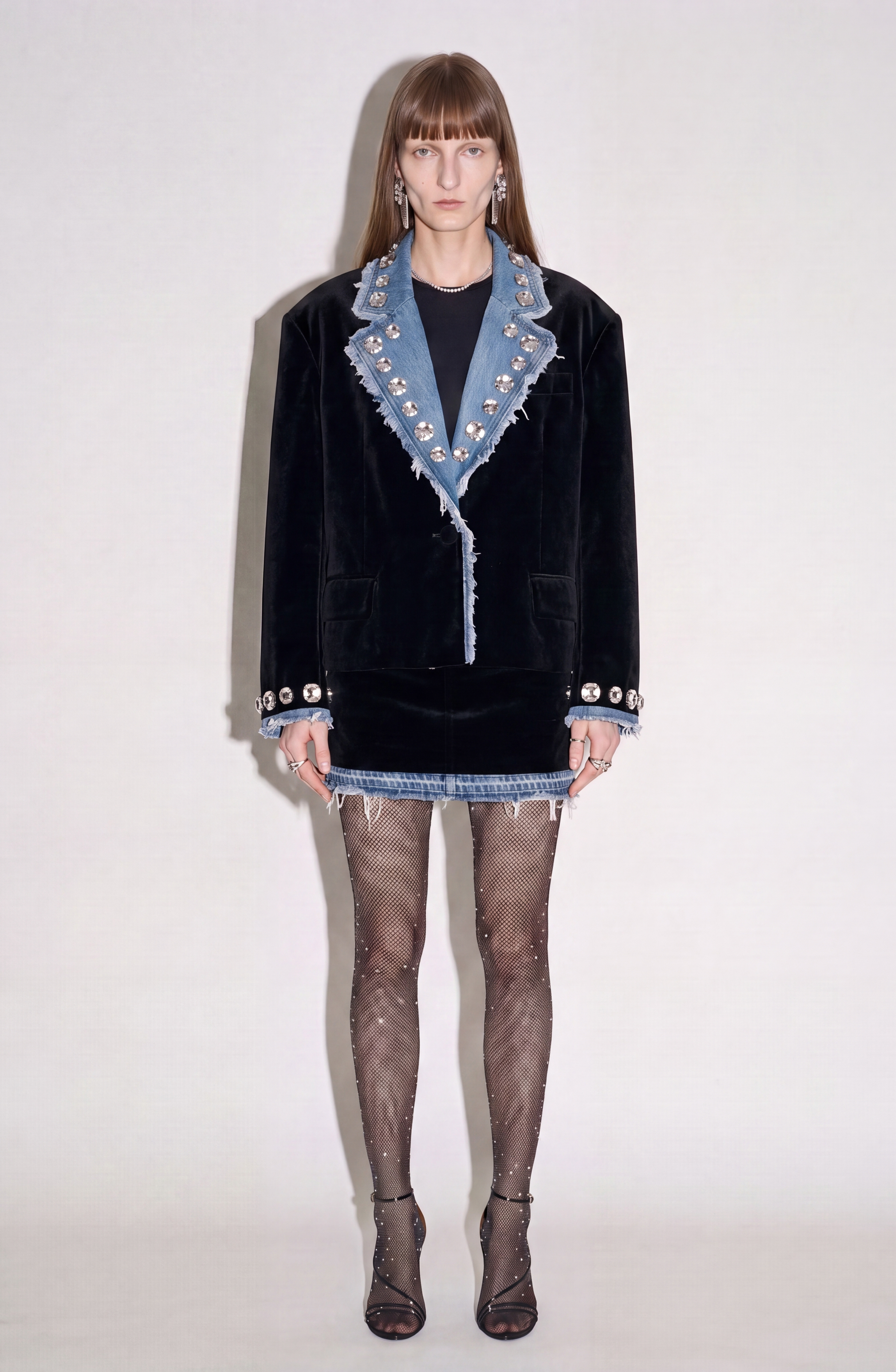 A woman stands against a plain background wearing the LOULOU Velvet And Denim Mini Skirt, which features a black velvet body and a denim hem. She pairs it with a matching black velvet blazer with denim accents, sheer polka-dot tights, and open-toe heels. Her straight brown hair is styled with bangs.