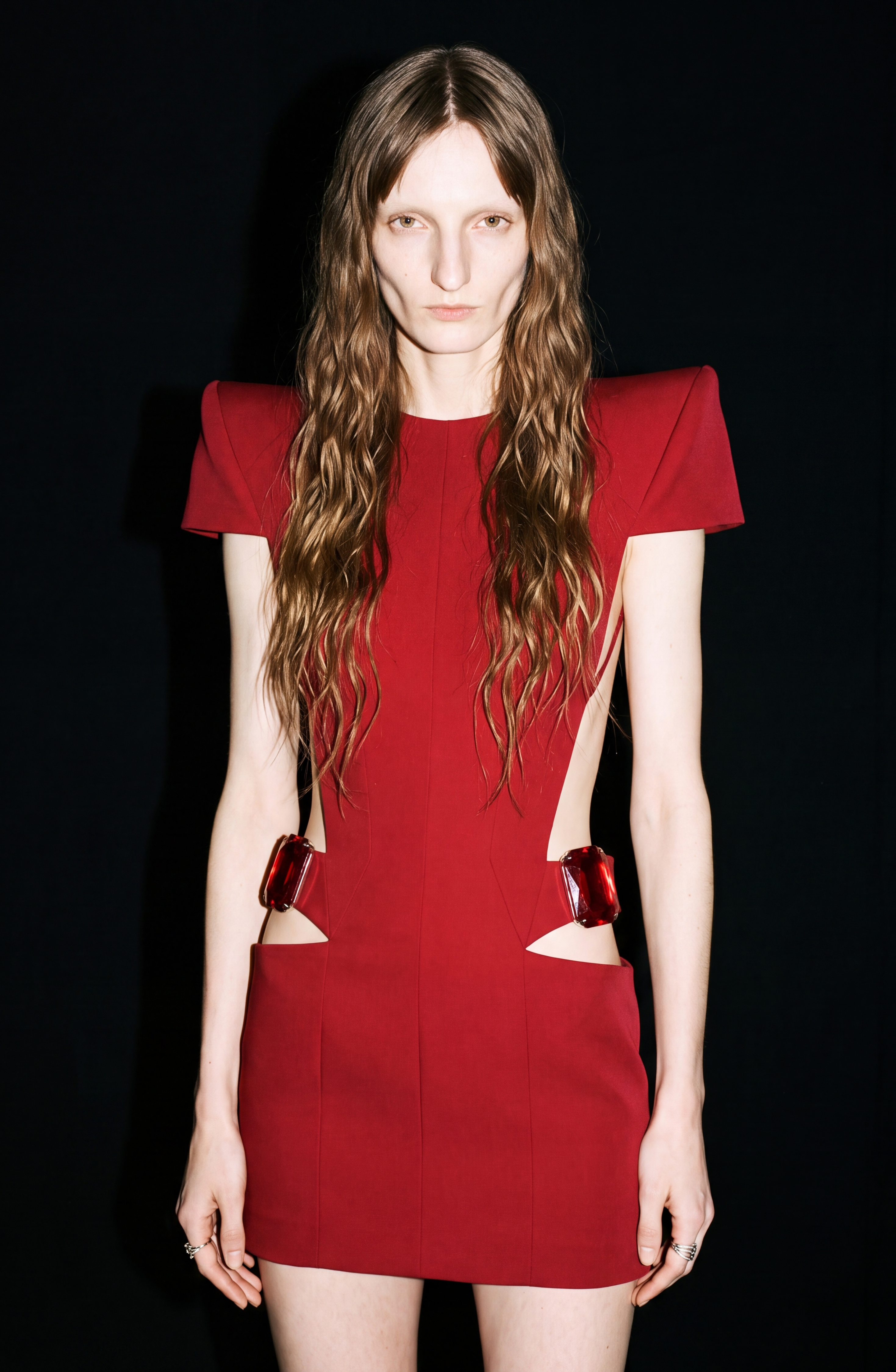 A person with long wavy hair wears the LOULOU Crystal-Embellished Cutout Mini Dress, a sleeveless, structured ruby red mini dress with side cutouts and padded shoulders, standing against a black background.
