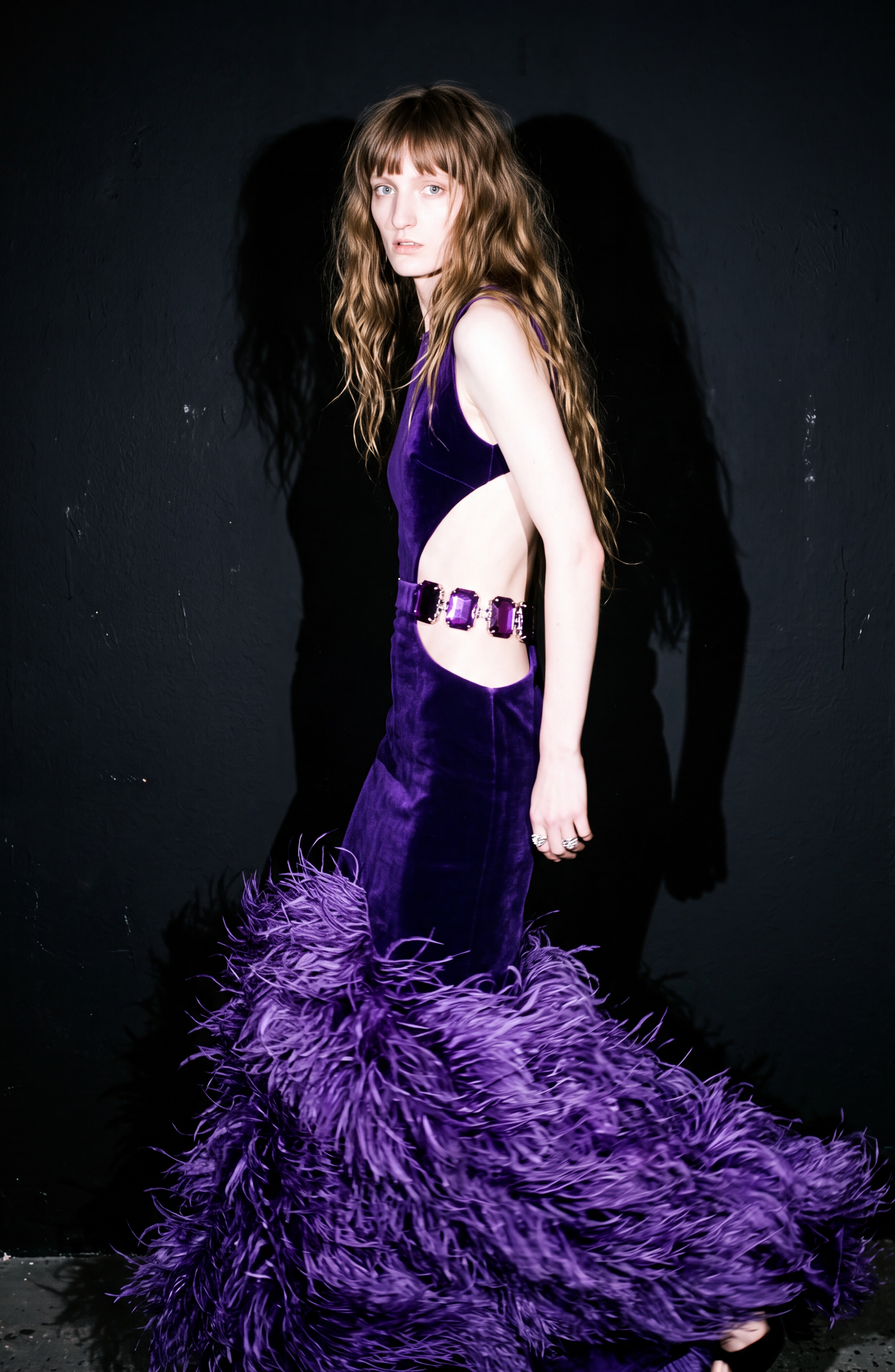 Dressed in the LOULOU Velvet Cutout Feather Gown, a figure stands against a dark background, the gown’s purple velvet and feather-trimmed skirt softly catching the light as their silhouette casts a shadow on the wall.