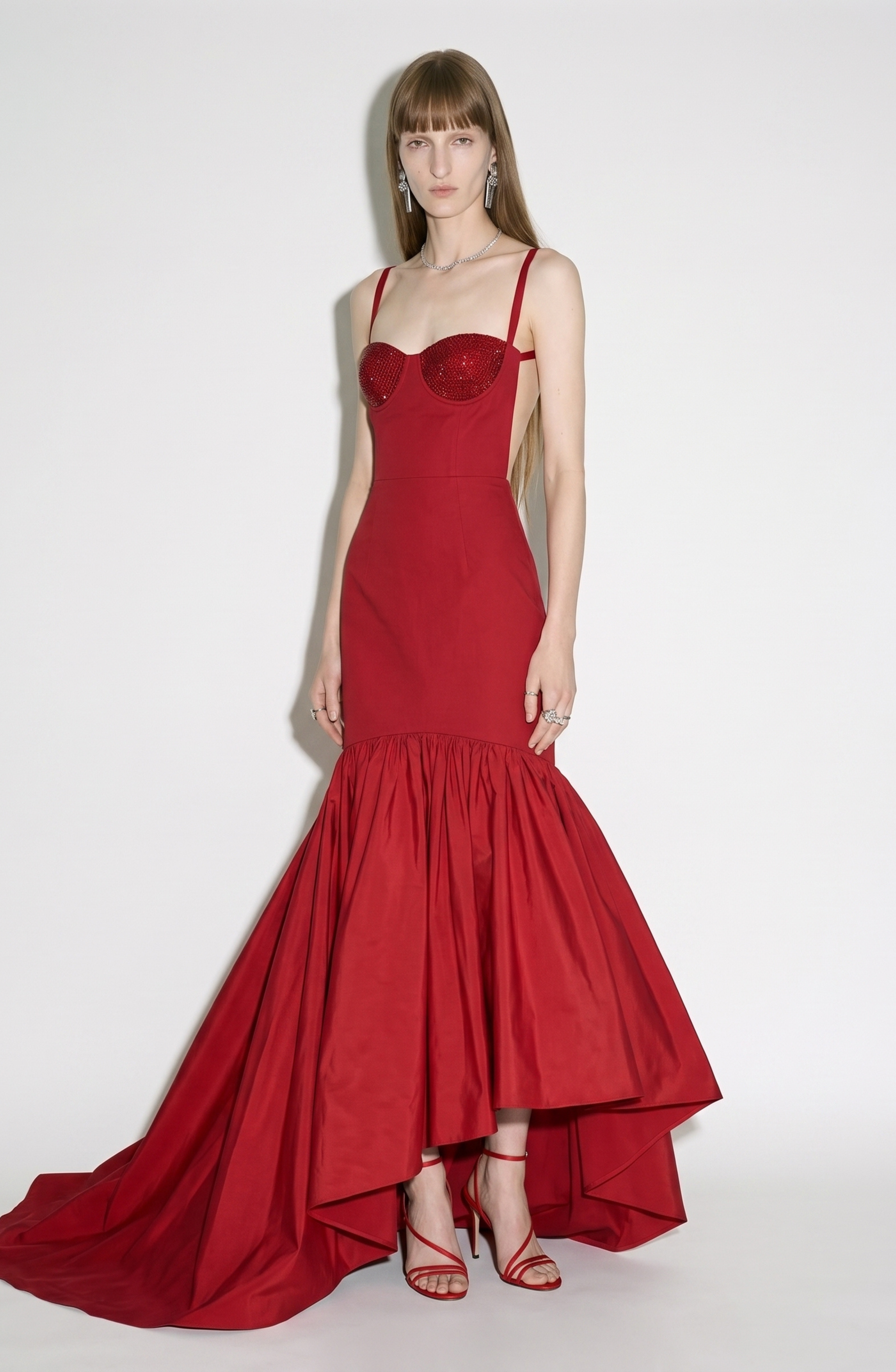 A person poses against a plain background wearing the LOULOU Crystal-Embellished Bustier Gown—a ruby red, strapless, mermaid-style dress featuring a crystal-adorned bodice and ruffled hem—paired with red high heels and long straight hair.