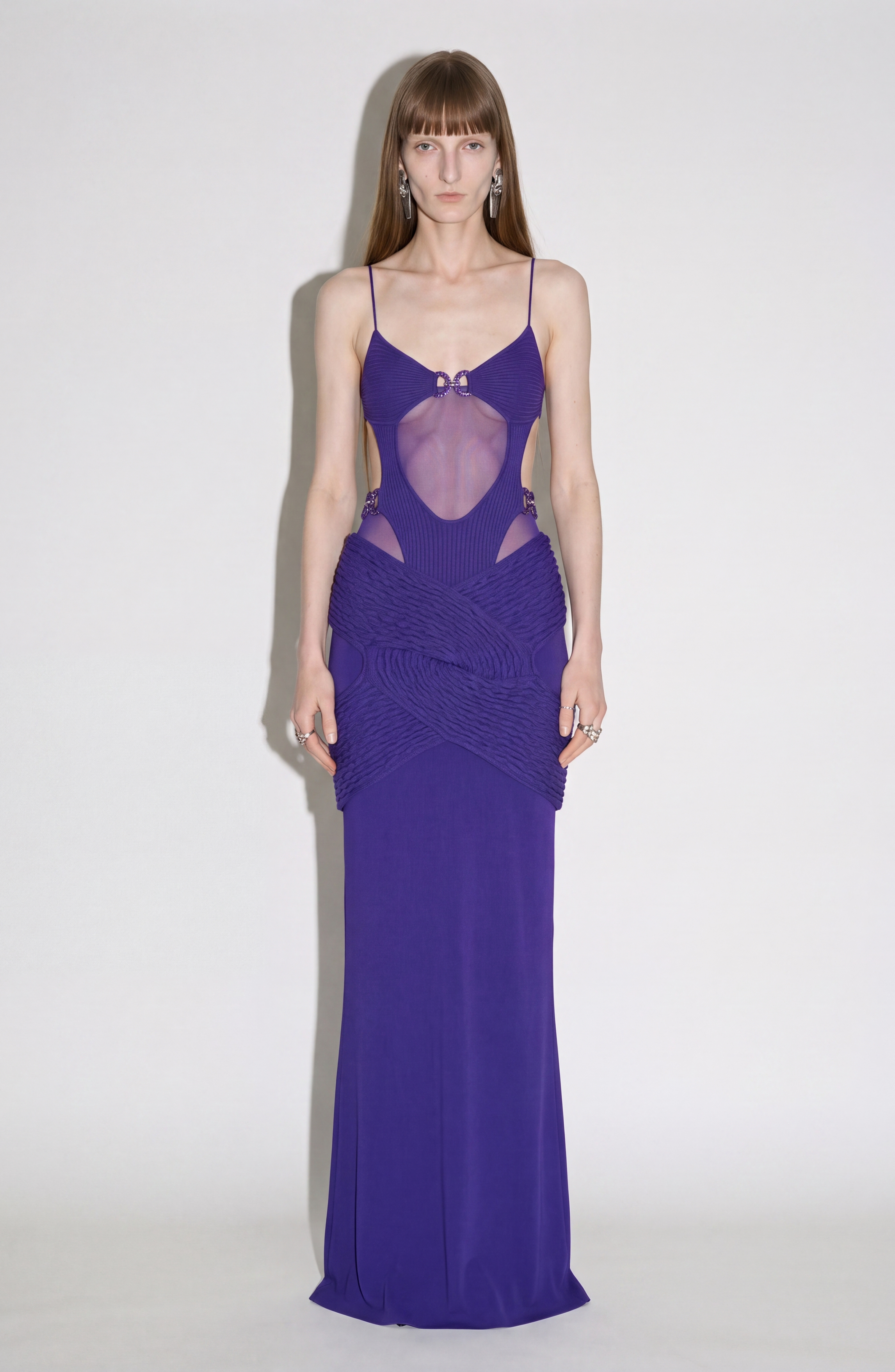 A person stands against a white background wearing the LOULOU Draped Cut-Out Knit Gown, a long, sleeveless maxi dress in deep purple with thin straps, cut-out details, and a sheer mesh front panel.
