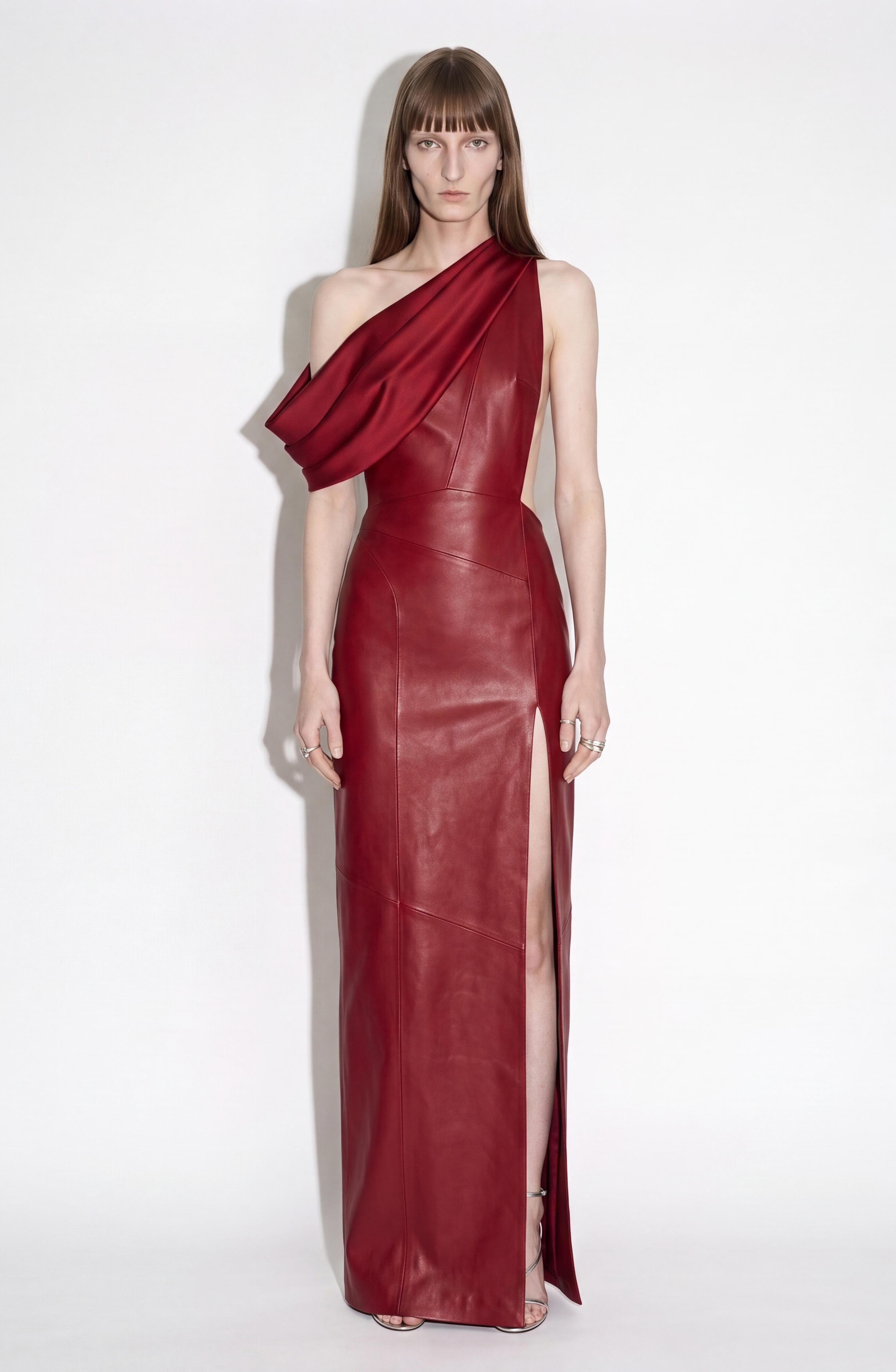 A person stands against a white background wearing the LOULOU One-Shoulder Leather Maxi Dress in oxblood, featuring a high slit and elegant draped detailing.