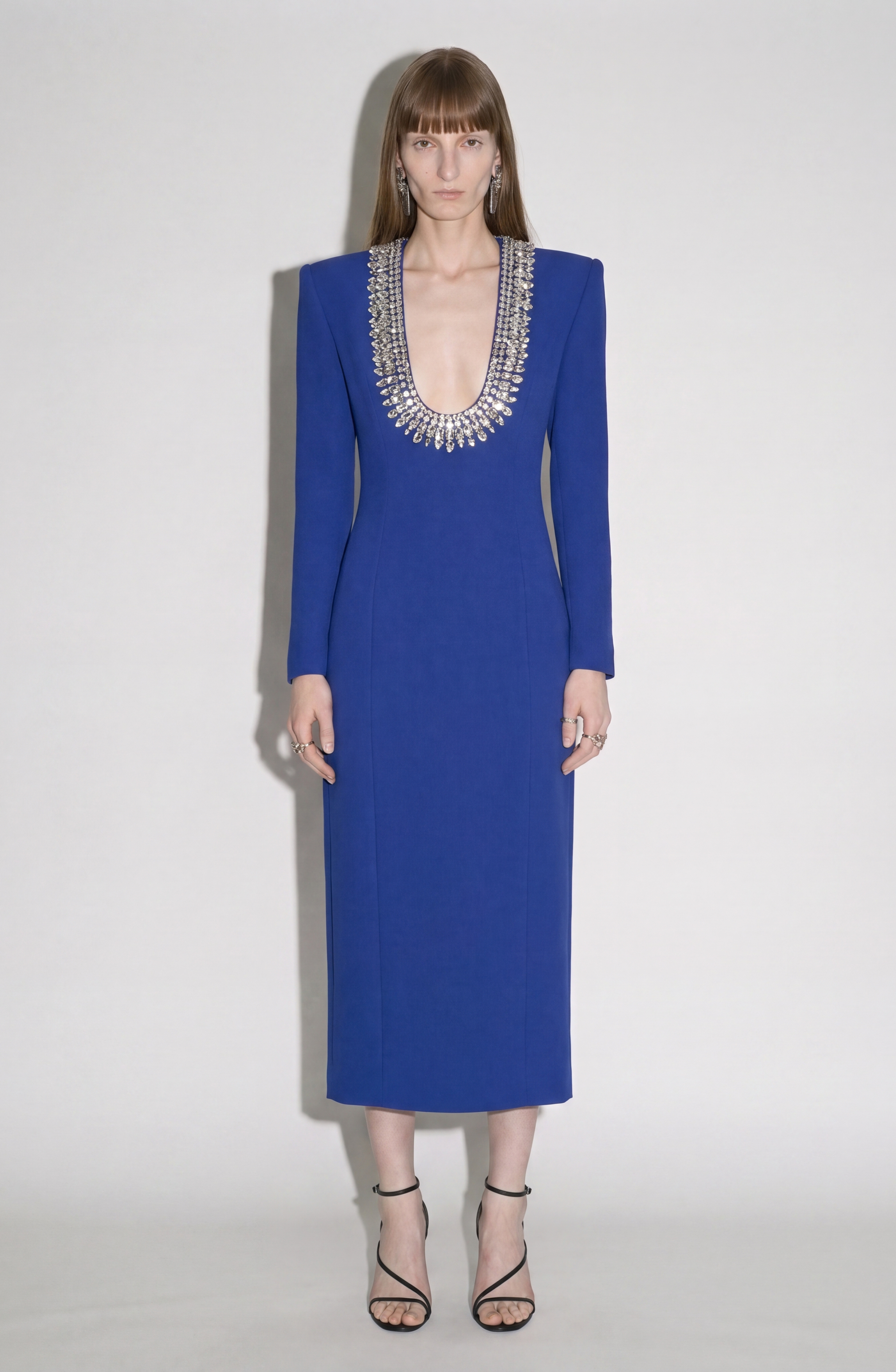 Wearing the LOULOU Crystal U-Neck Midi Dress in royal blue—with long sleeves, padded shoulders, and a deep, crystal-embellished neckline—a person pairs it with black strappy heels and stands against a plain white background.