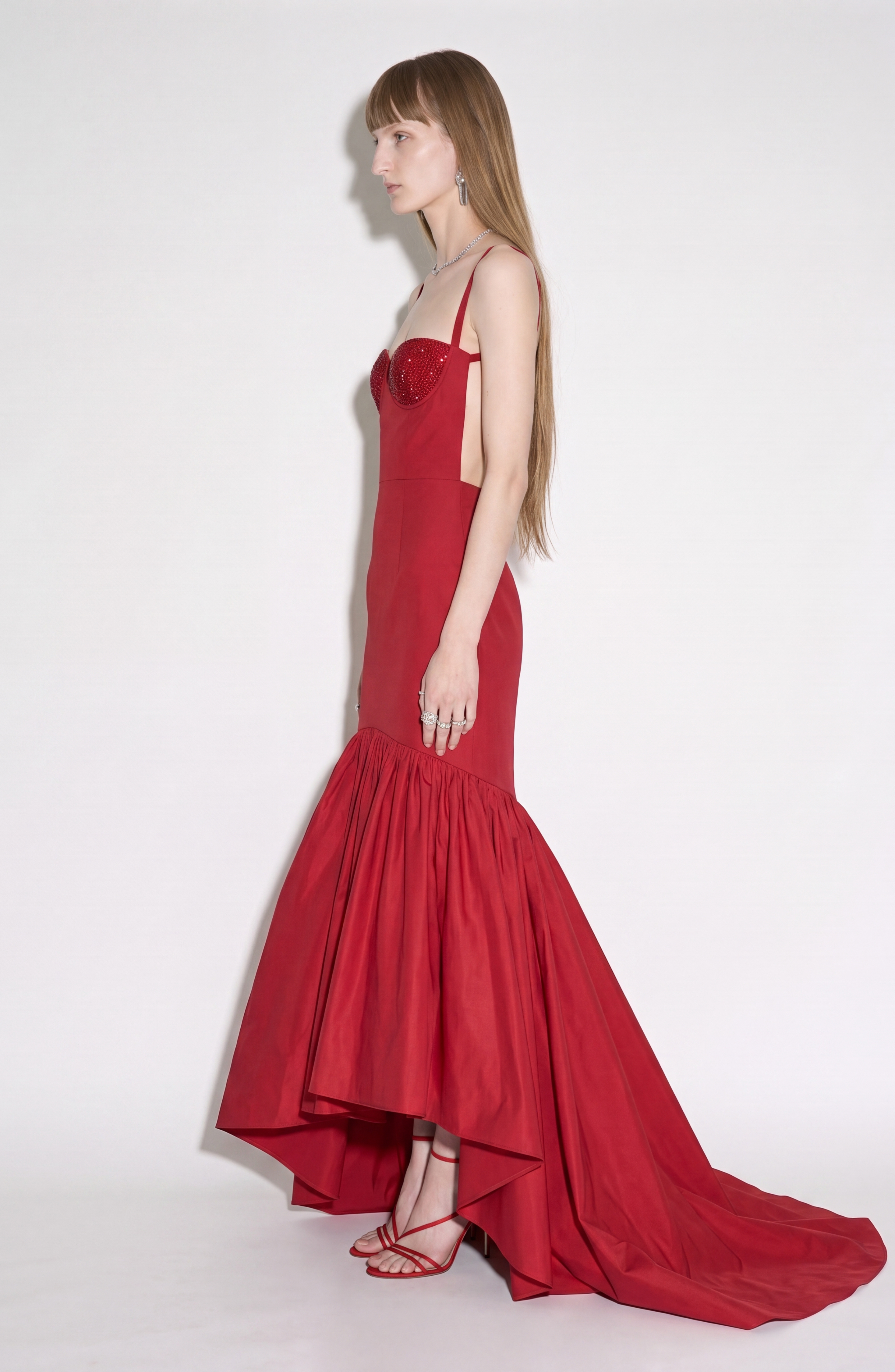 A woman stands in profile against a plain white background, wearing the LOULOU Crystal-Embellished Bustier Gown—featuring a fitted bodice adorned with sparkling crystals, a flared ruby red skirt, and paired with red strappy heels.