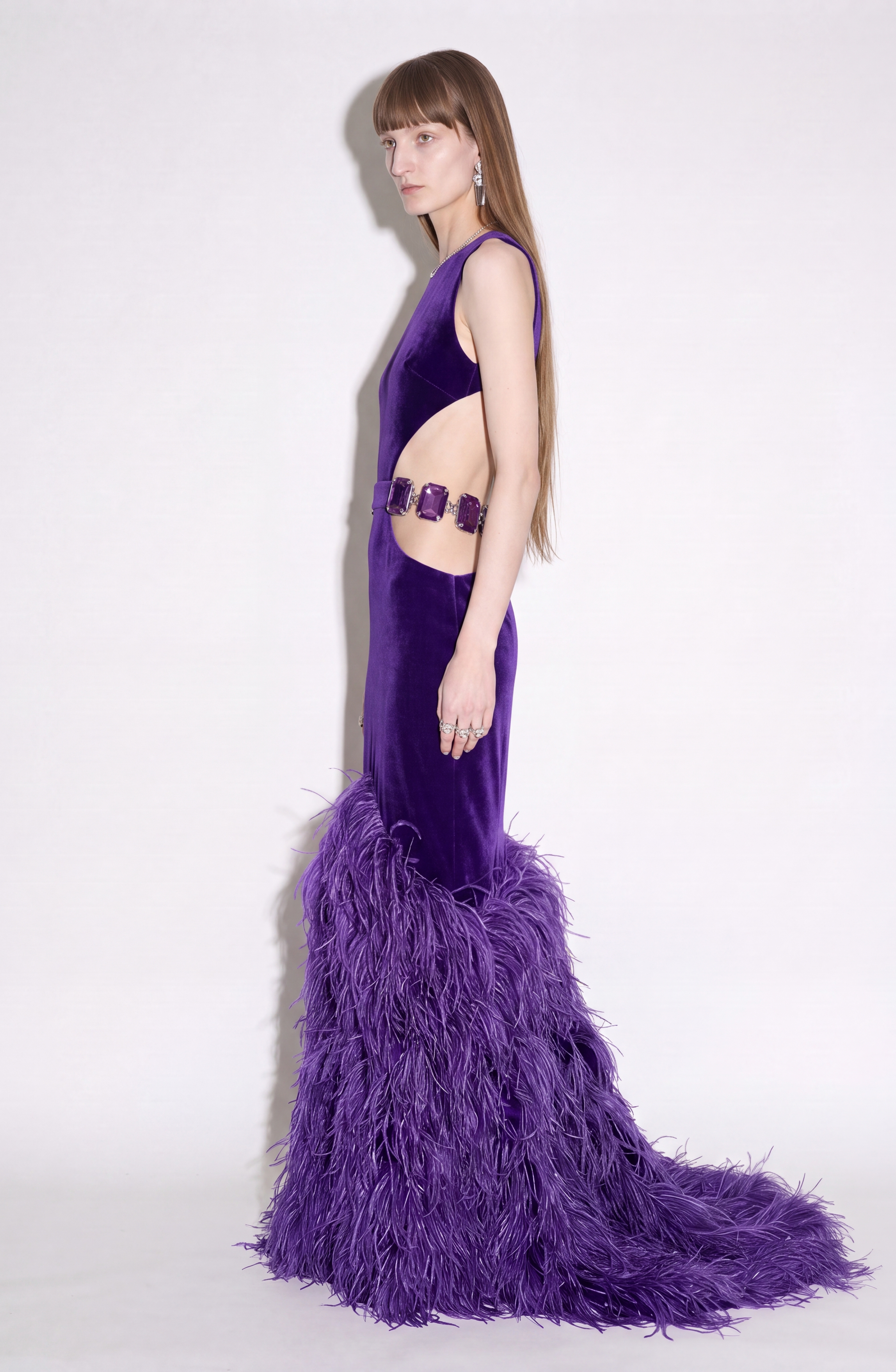 A woman with long straight hair poses in profile against a plain white background, wearing the LOULOU Velvet Cutout Feather Gown featuring side cut-outs and feathered details at the hem.