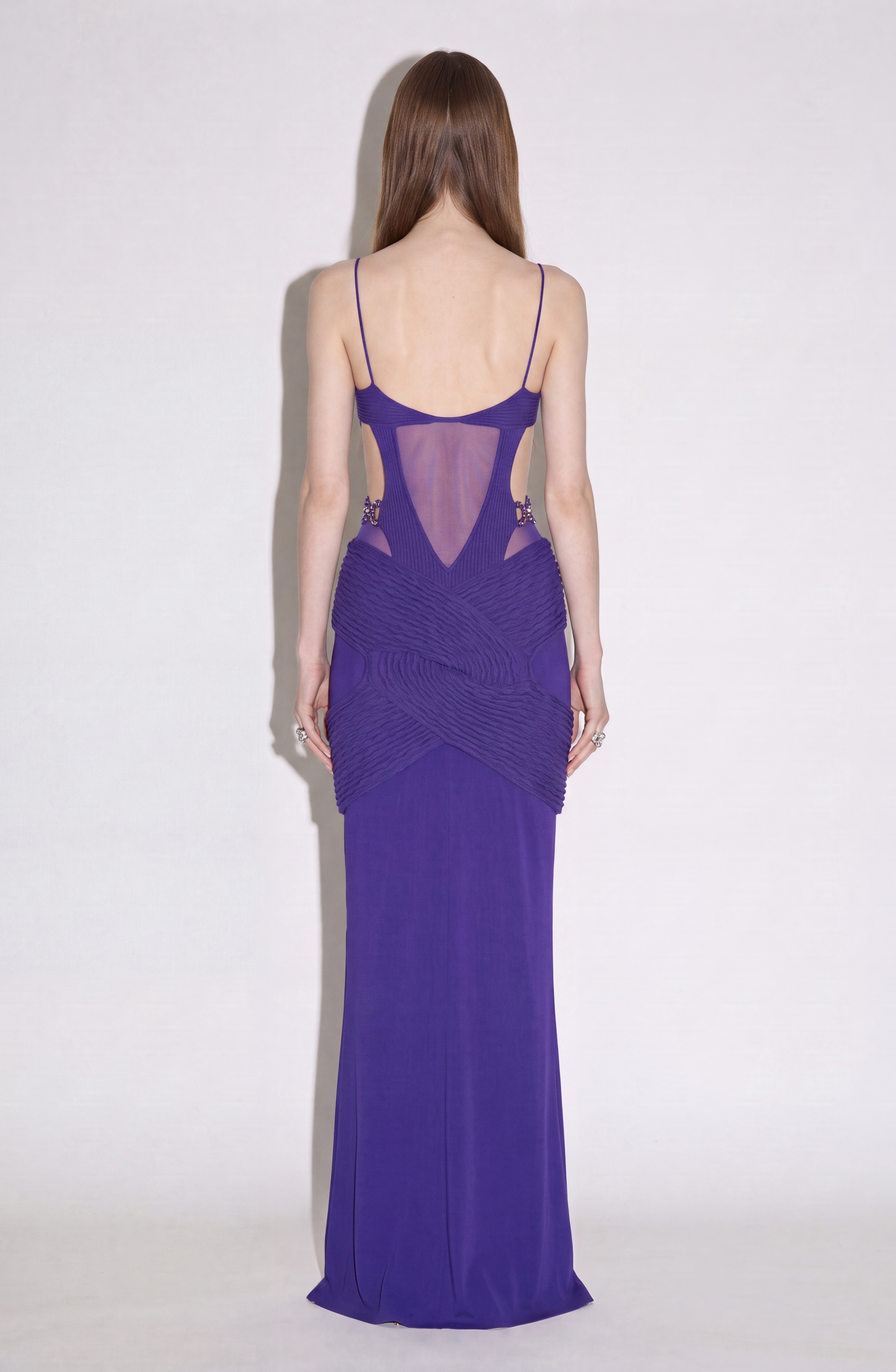 A person with long brown hair, shown from the back and facing a white wall, is wearing the LOULOU Draped Cut-Out Knit Gown—a sleeveless purple maxi dress featuring sheer mesh and textured details.