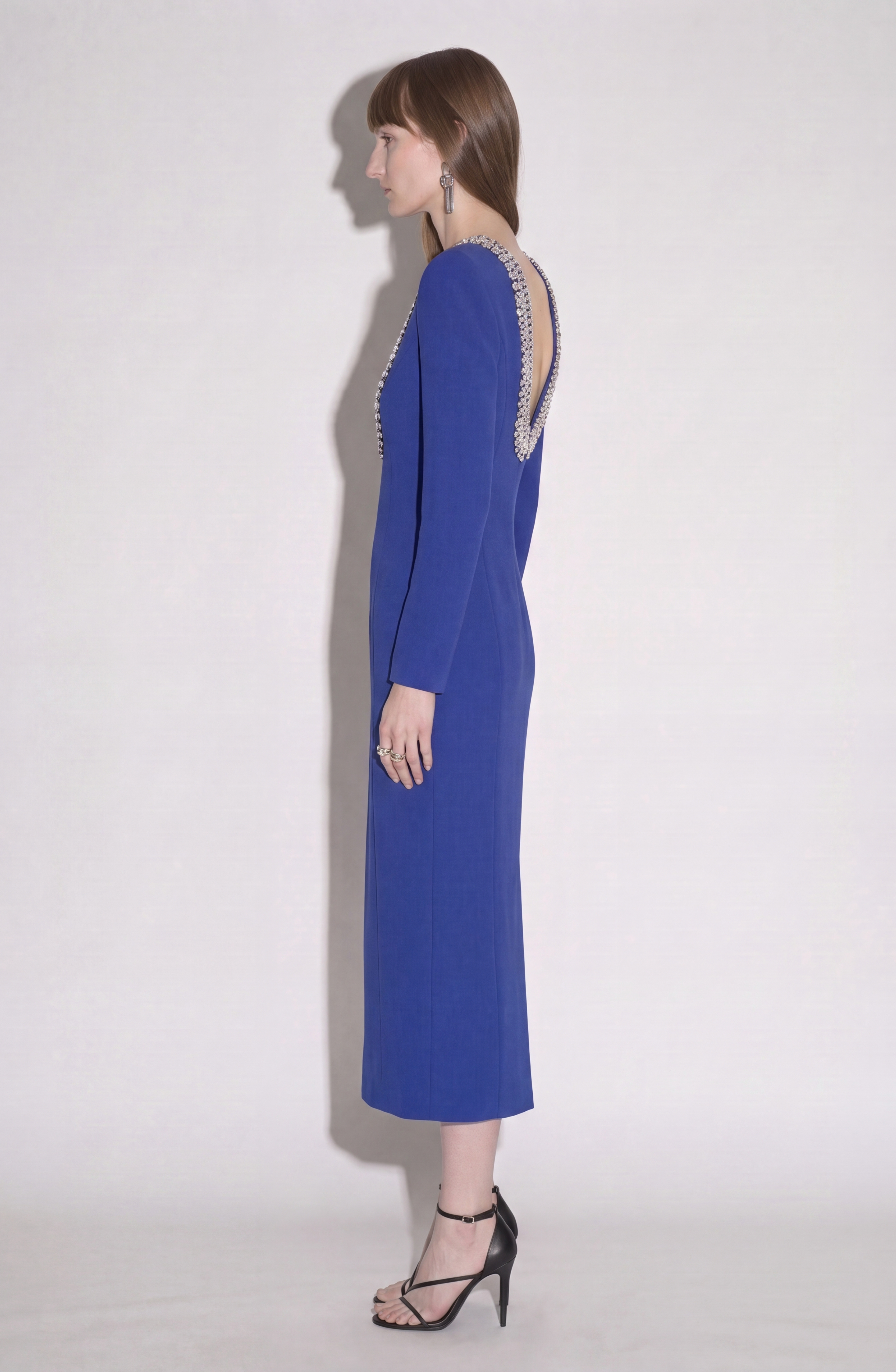 A woman wearing the LOULOU Crystal U-Neck Midi Dress, featuring royal blue fabric, long sleeves with padded shoulders, a deep V-cut back, and beaded trim, stands in profile against a plain light background, accessorized with black strappy heels and long earrings.