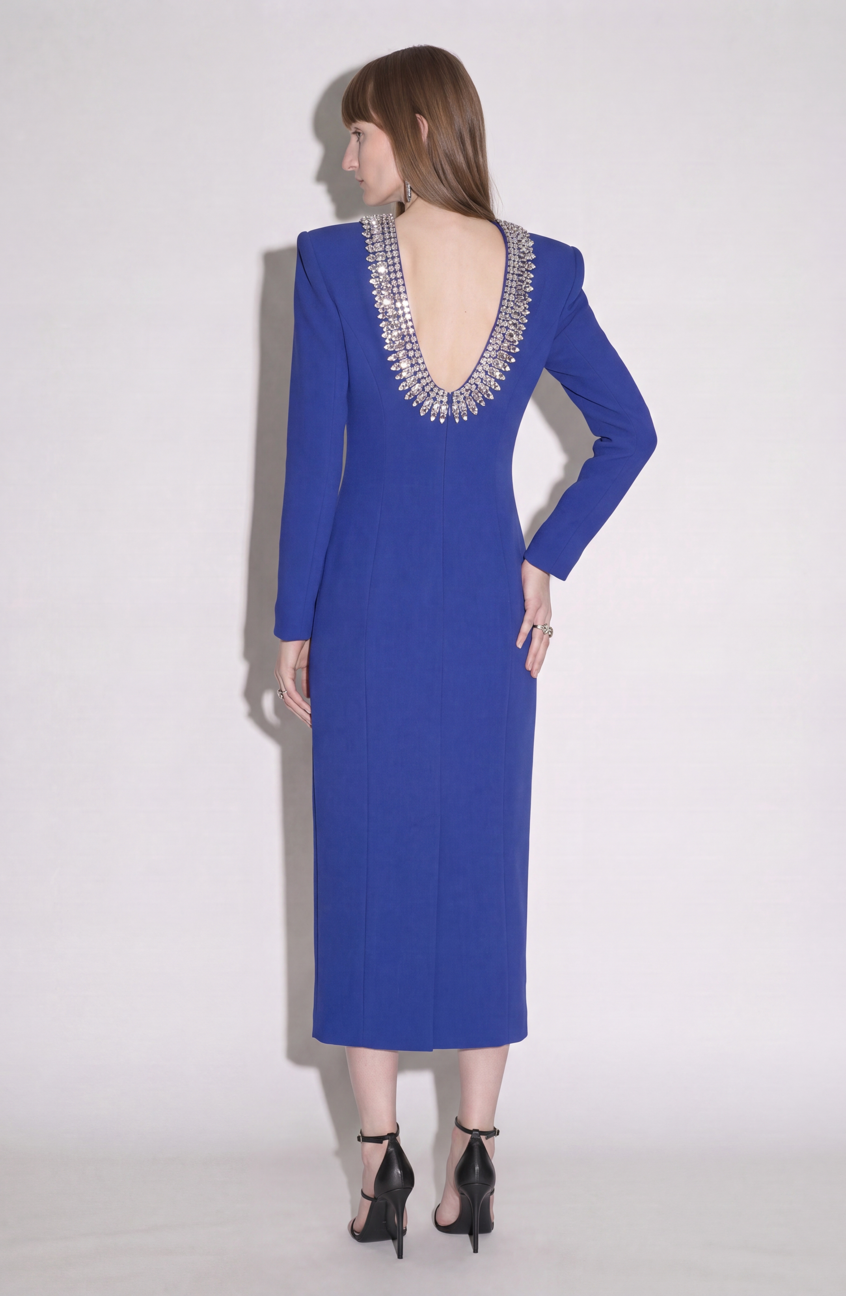 A woman stands facing away with one hand on her hip, wearing the LOULOU Crystal U-Neck Midi Dress in blue. The dress features long sleeves, padded shoulders, a dramatic backless design, and silver crystal embellishments at the neckline. She completes the look with black heels.