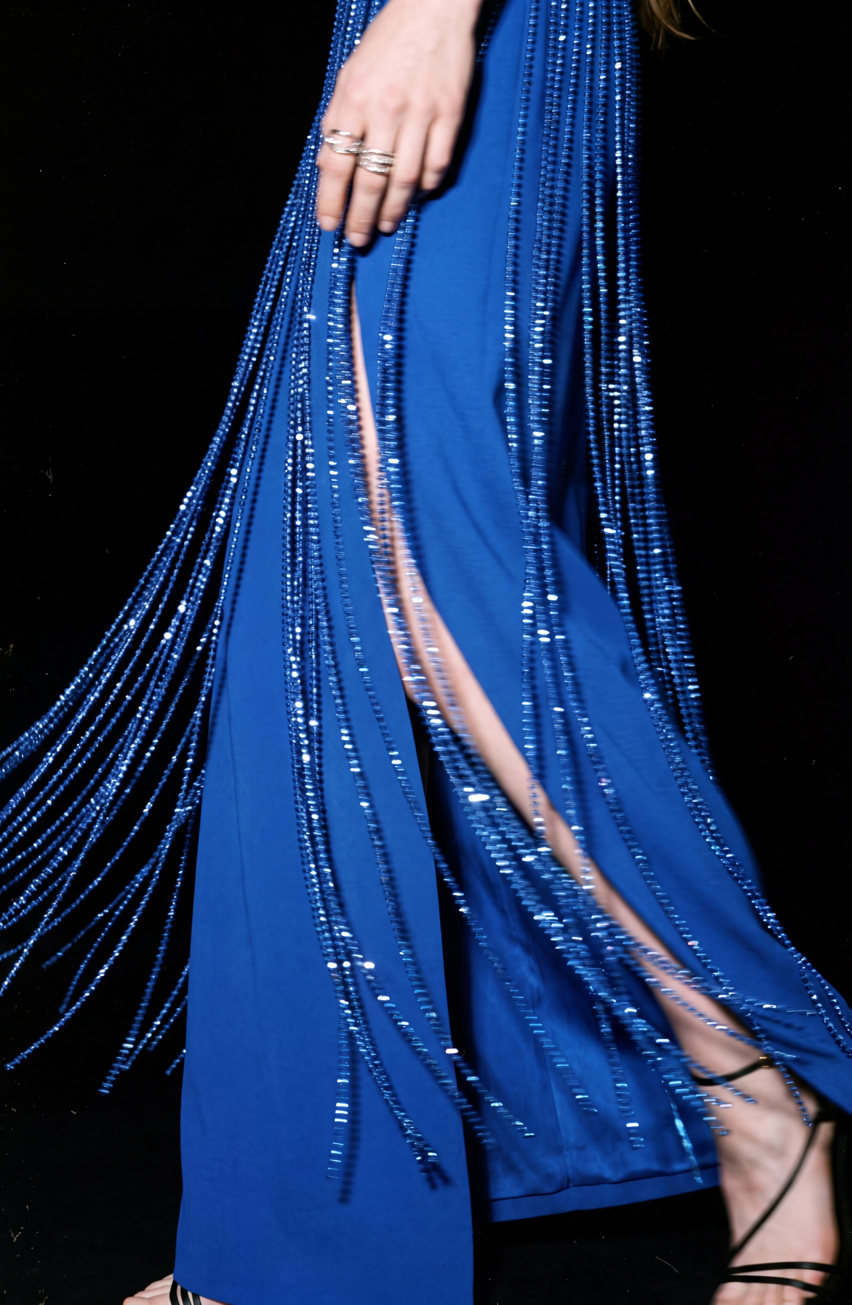 A person wearing the LOULOU Cobalt Crystal Fringe Maxi Dress, featuring shimmering beaded fringe reminiscent of a crystal-fringed skirt and a thigh-high slit that reveals part of their leg. Their hand adorned with rings and strappy black sandals are also visible.