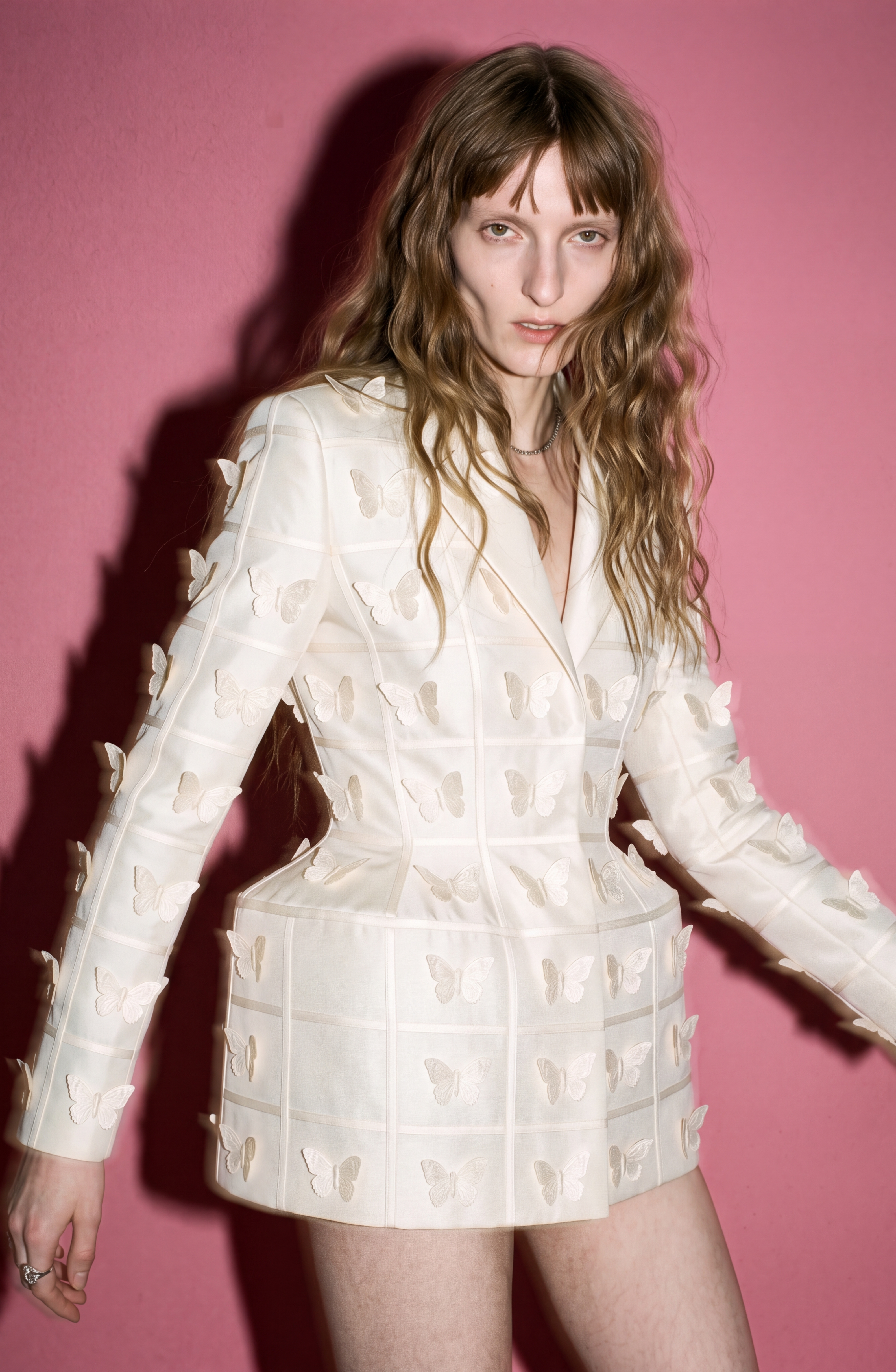 A person with long wavy hair wears the LOULOU Embellished Silk Basque Blazer Dress in ivory, featuring delicate butterfly appliqués, while standing against a solid pink background.