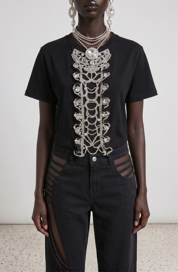 A person wears the No Notes T-Shirt by LOULOU—black cotton with hand-applied glass crystals in a baroque-inspired necklace and earring motif—paired with black mesh cut-out pants. The background is plain and neutral.