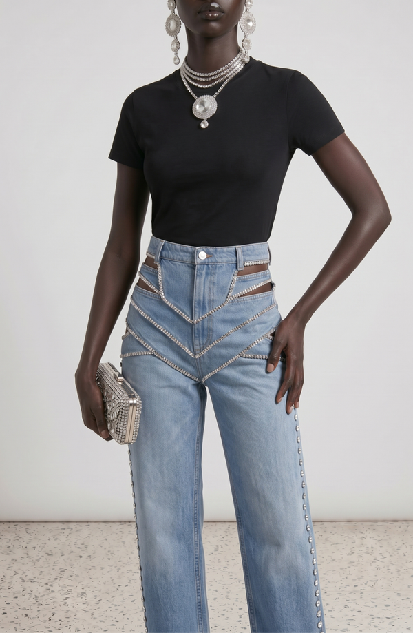 A person wearing a black t-shirt, LOULOU Runaway Jeans with chains and cut-out details, statement jewelry, and holding a studded clutch stands against a plain background.