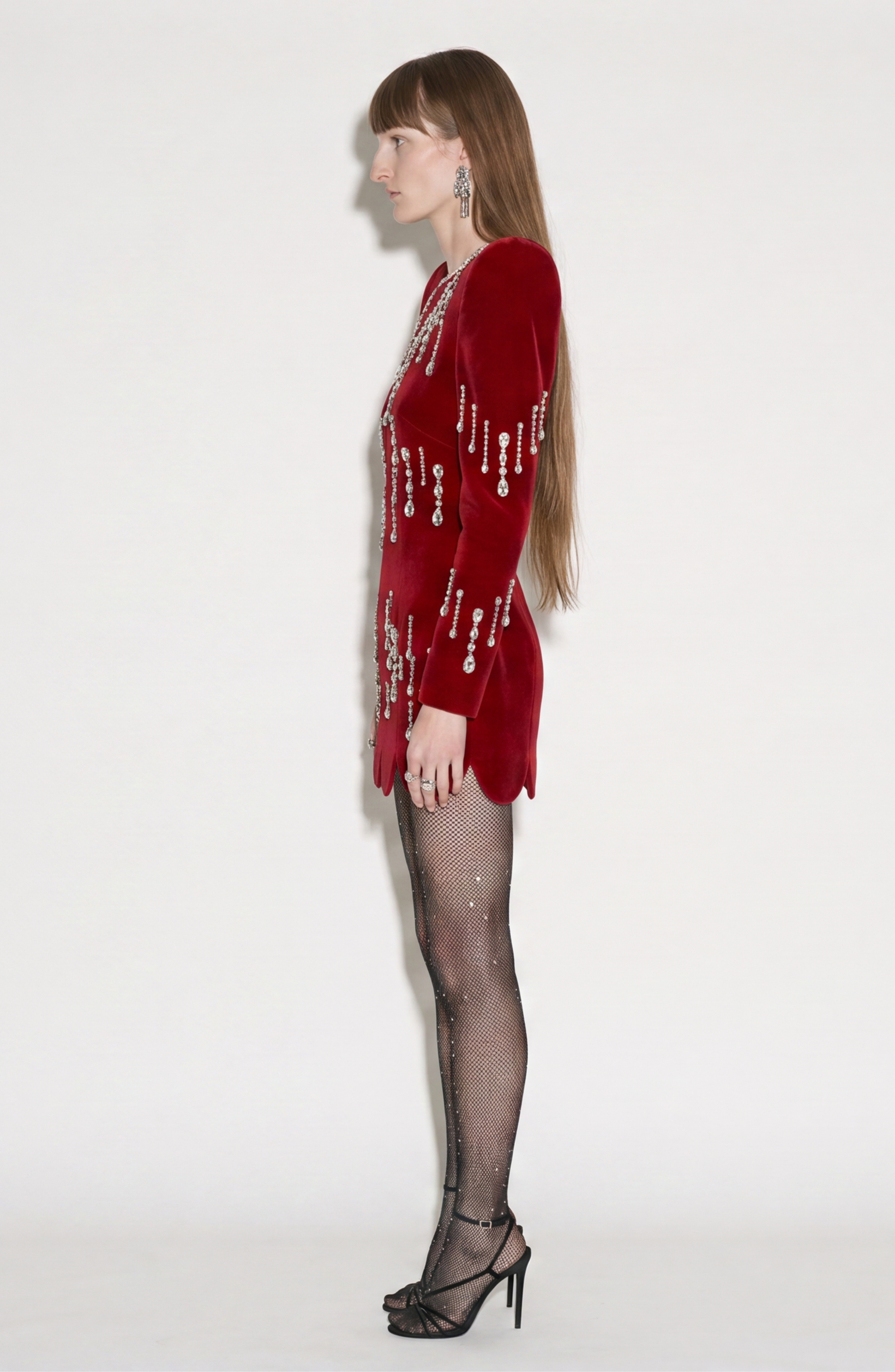 A person with long straight hair stands in profile against a white background, wearing LOULOU’s Scarlet Velvet Mini Dress with sculptural shoulders, paired with black fishnet tights, black heels, and large earrings.
