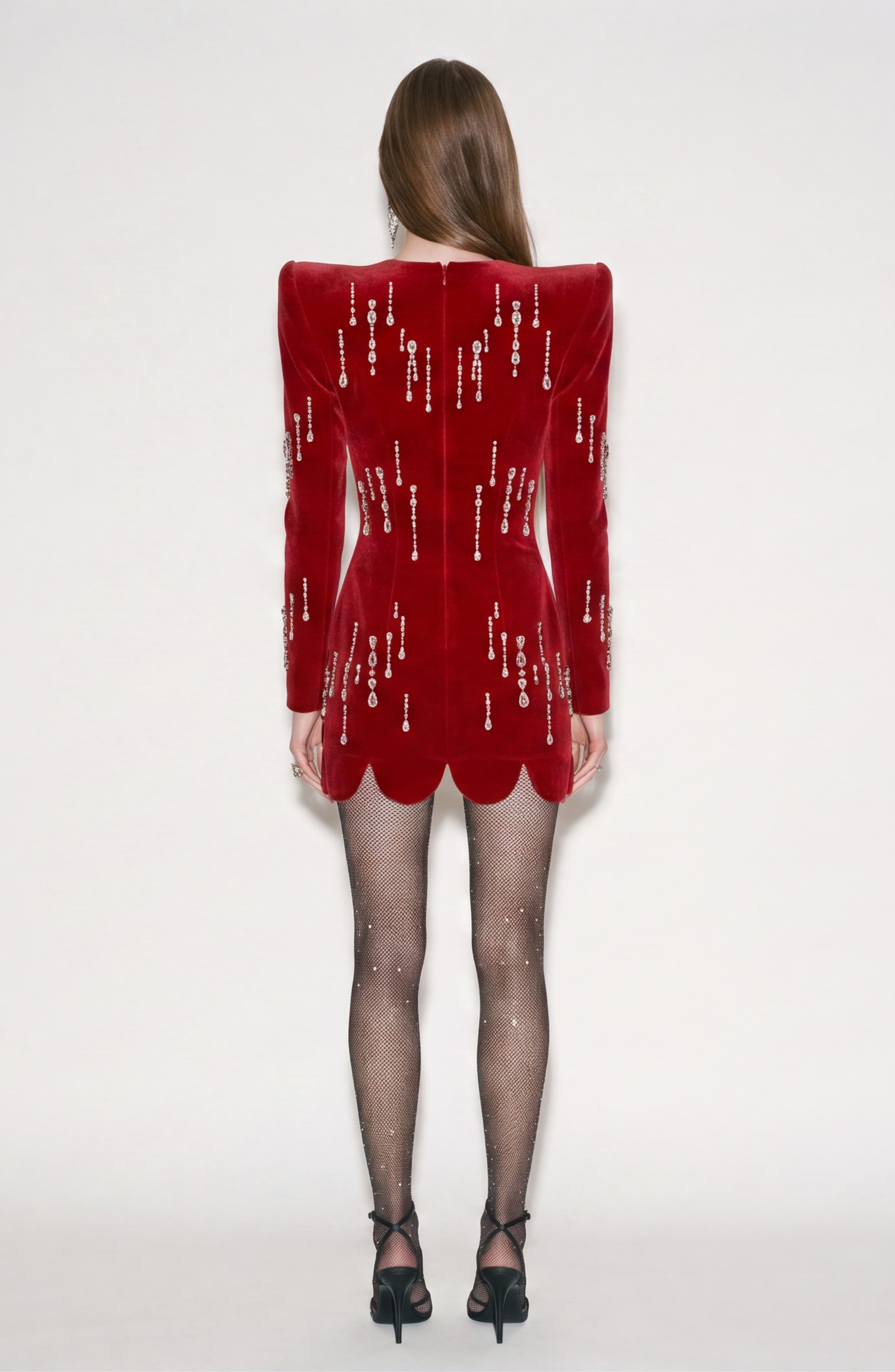 A woman faces away, wearing the LOULOU Scarlet Velvet Mini Dress with sculptural shoulders, scalloped hem, and sparkly text details, styled with sheer black tights and black heels against a plain white background.