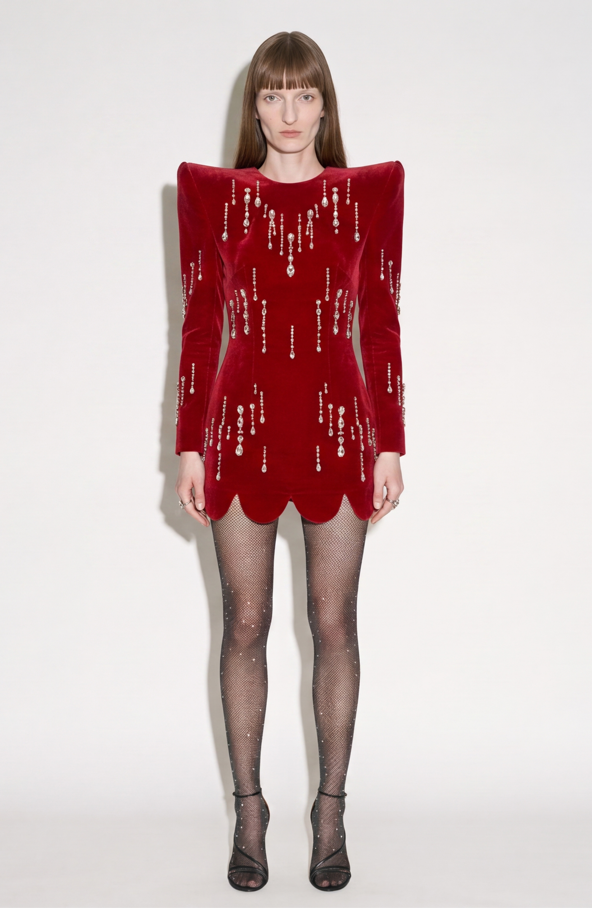 A woman poses against a white background in the LOULOU Scarlet Velvet Mini Dress, featuring sculptural shoulders, scalloped hem, and glass crystal embellishments. She completes the look with sheer sparkling black tights and black open-toe heels.