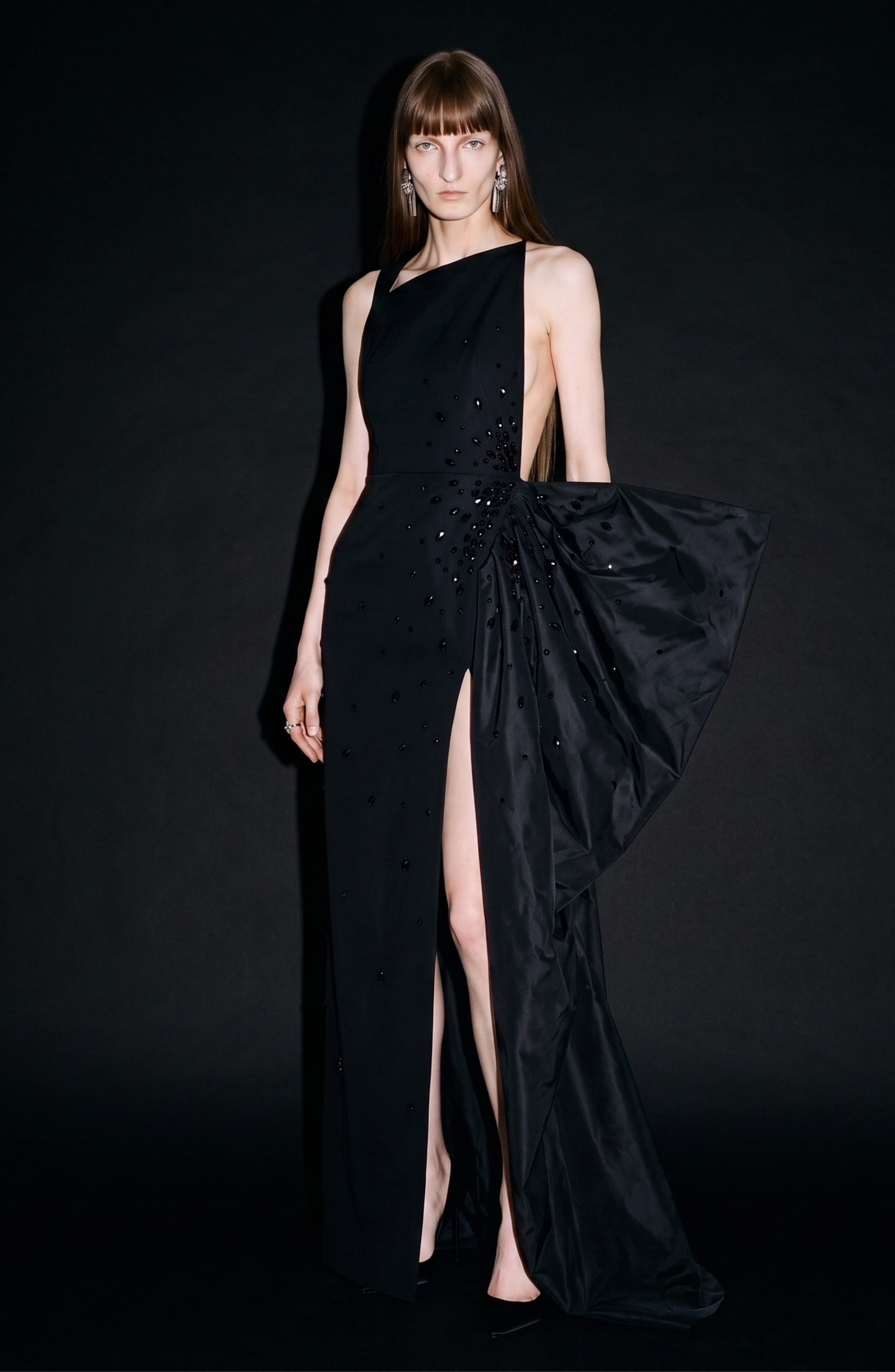 A person poses against a black background wearing the LOULOU Asymmetric Crystal Bow Gown, a sleeveless jet-black dress featuring an asymmetric neckline, sparkling crystal embellishments, and a dramatic draped side detail.
