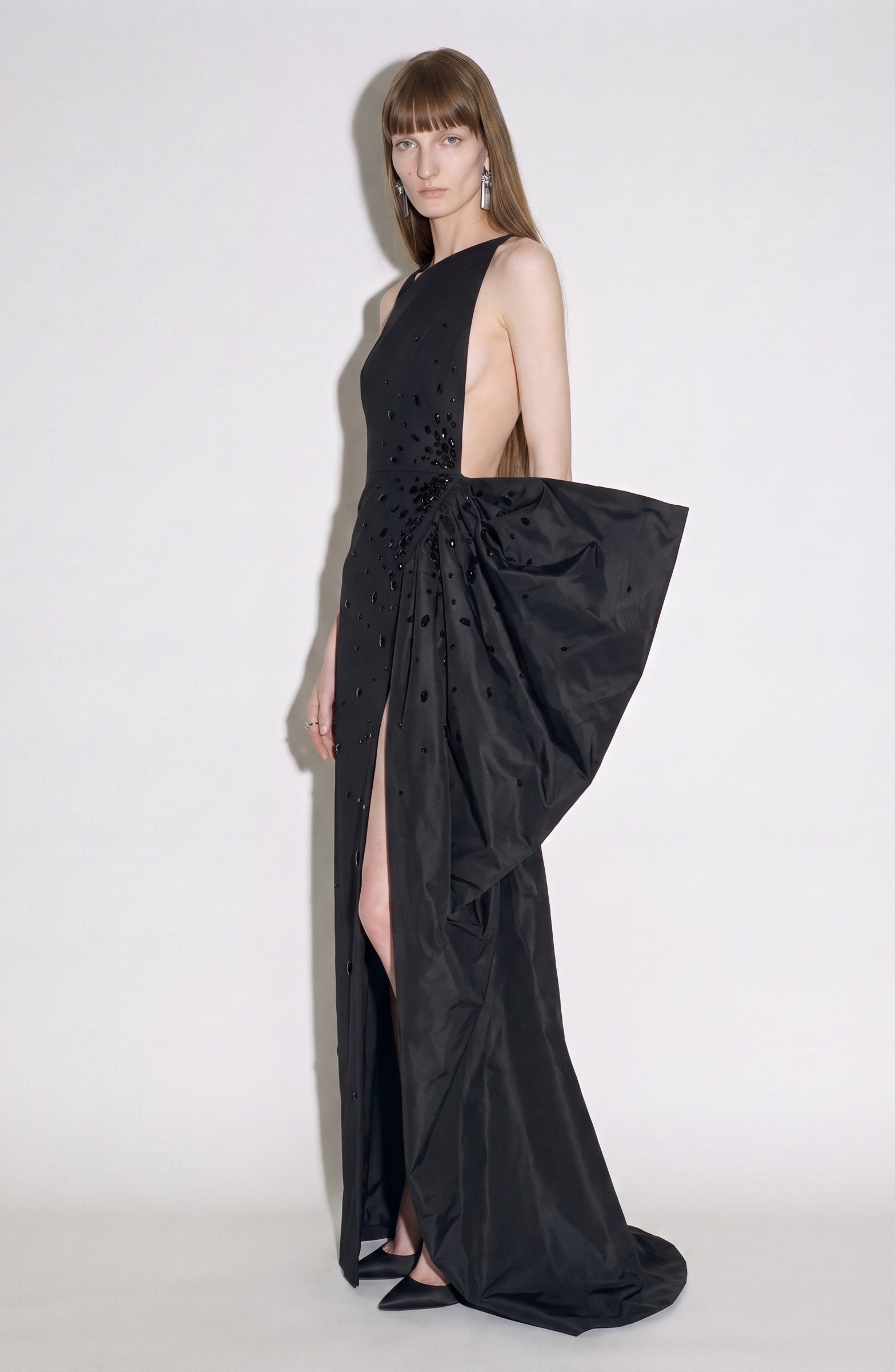 A woman poses against a plain white background wearing the LOULOU Asymmetrical Embellished Gown, featuring an asymmetrical neckline, side cutout, high slit, and an oversized bow detail. She has straight light brown hair and accessorizes with long earrings and black heels.