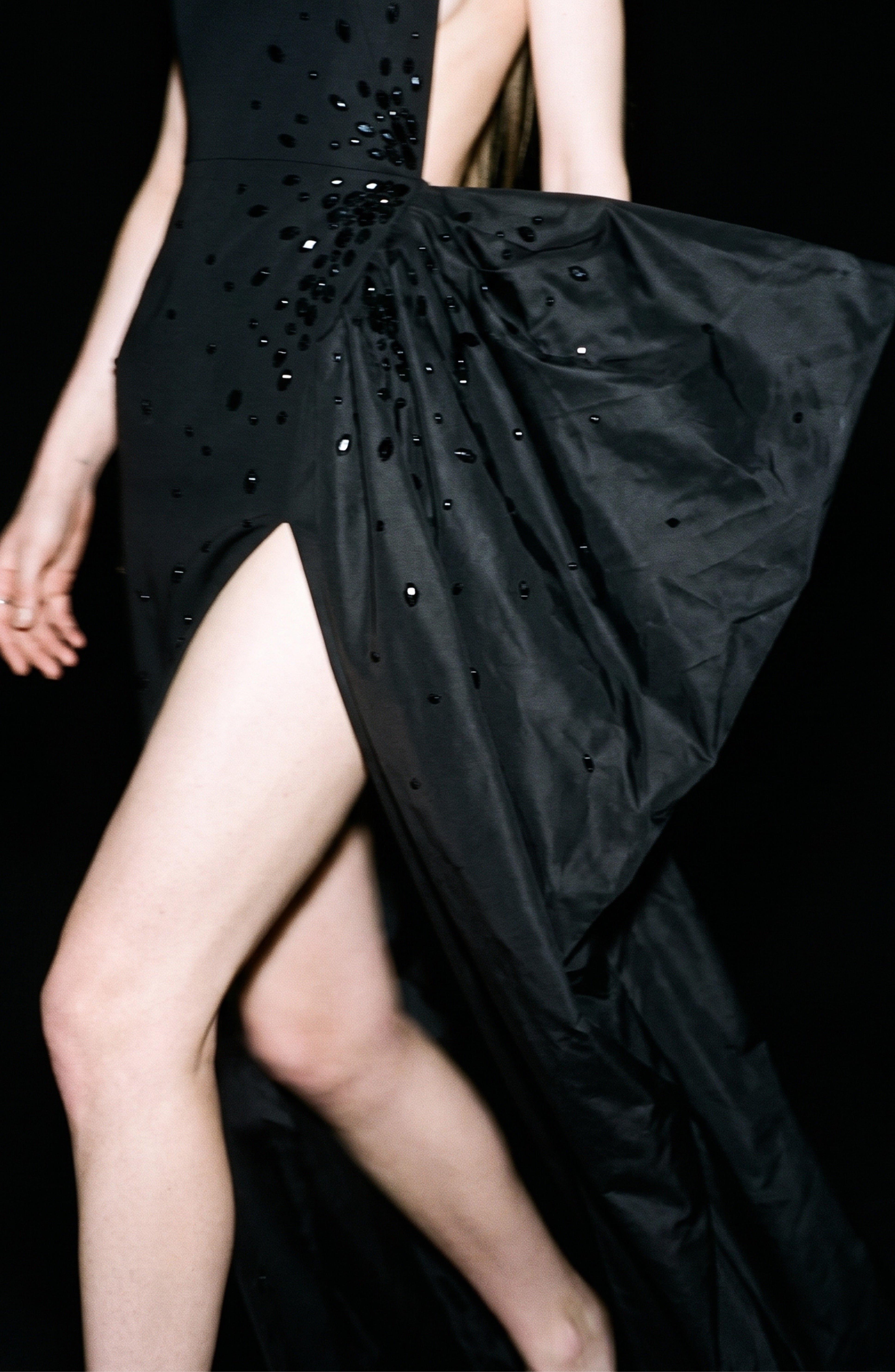 A model showcases the LOULOU Asymmetrical Embellished Gown, featuring an asymmetrical neckline and decorative black beads, while walking against a black background with one arm and leg visible.