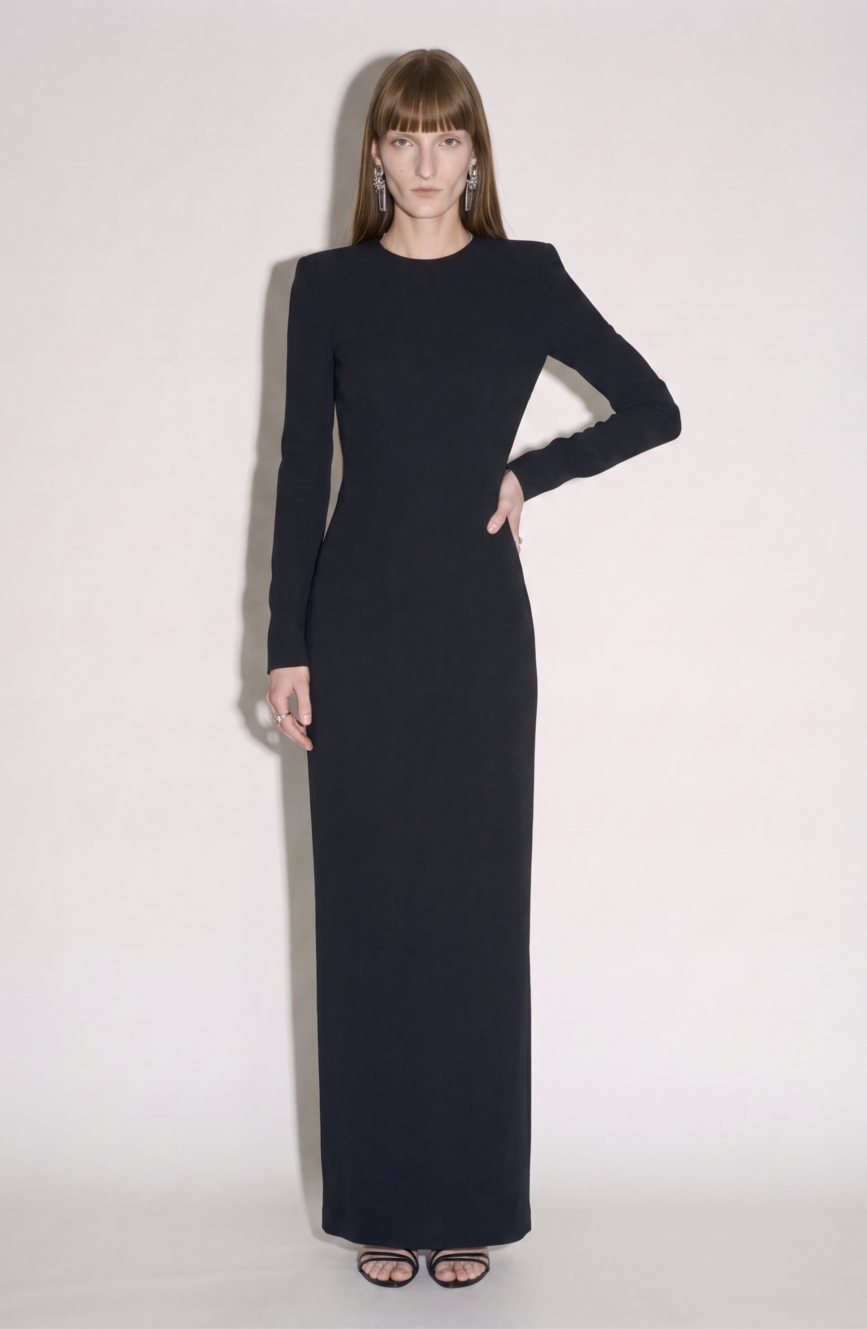A woman stands against a light background wearing the LOULOU Back Crepe Maxi Dress, a long, form-fitting black maxi dress with long sleeves, paired with black sandals. She has straight light brown hair with bangs and is accessorized with drop earrings.