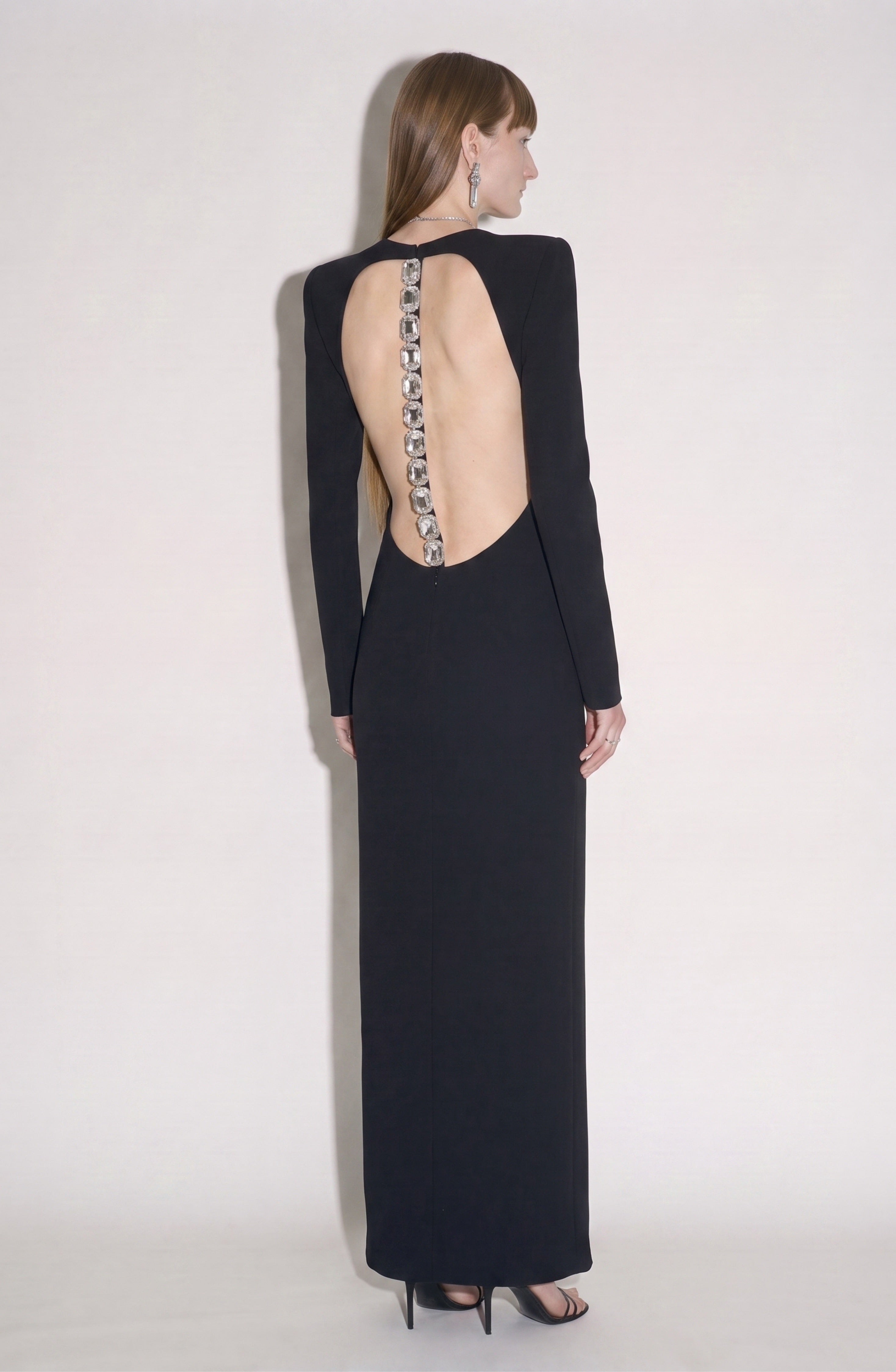 Model wearing the LOULOU Back Crepe Maxi Dress—an elegant, floor-length black dress featuring long sleeves and an open back highlighted by decorative silver square embellishments running down the spine—posed against a plain light background.